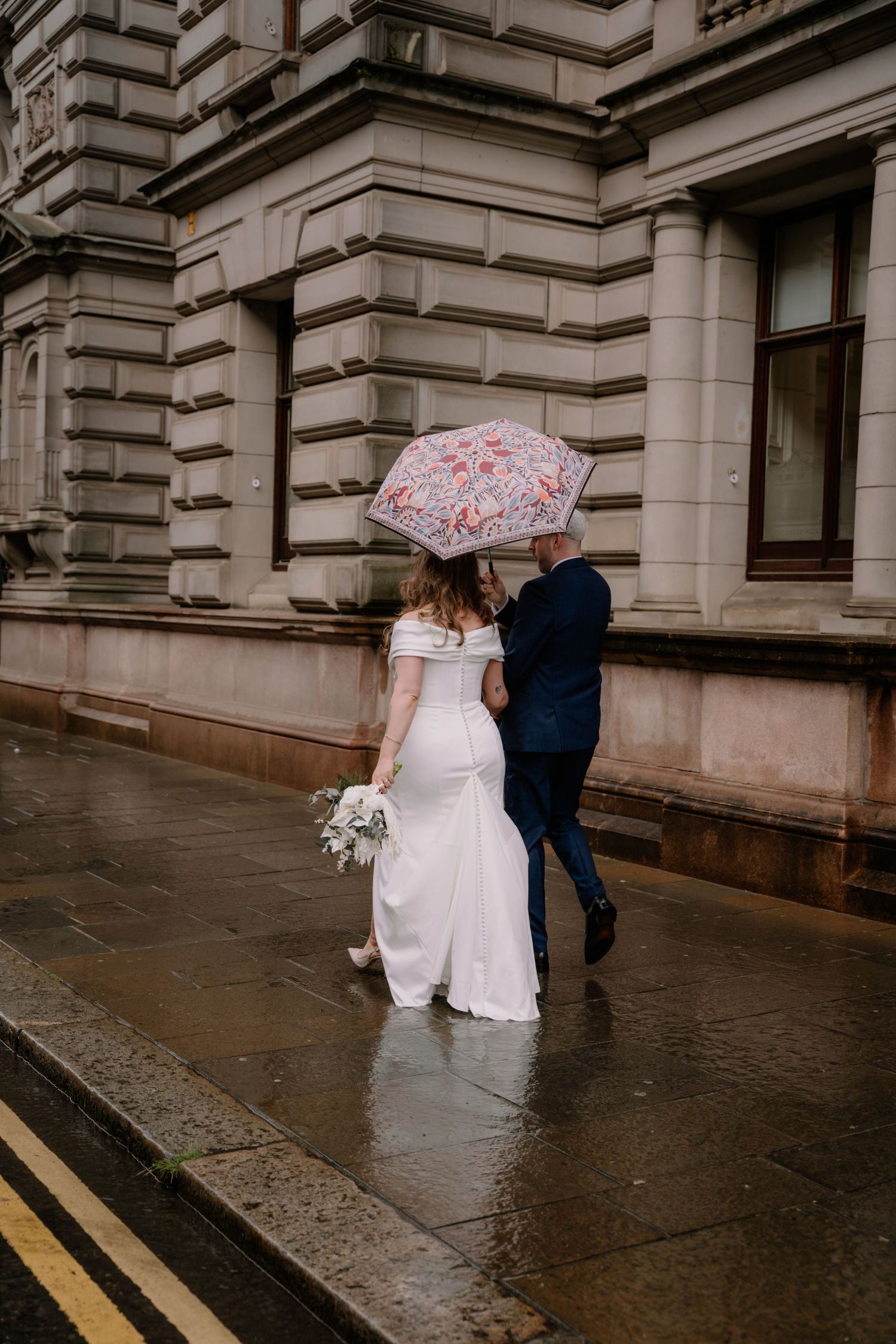 Bride and groom walking in rain under floral umbrella outside historic stone building on their wedding day.