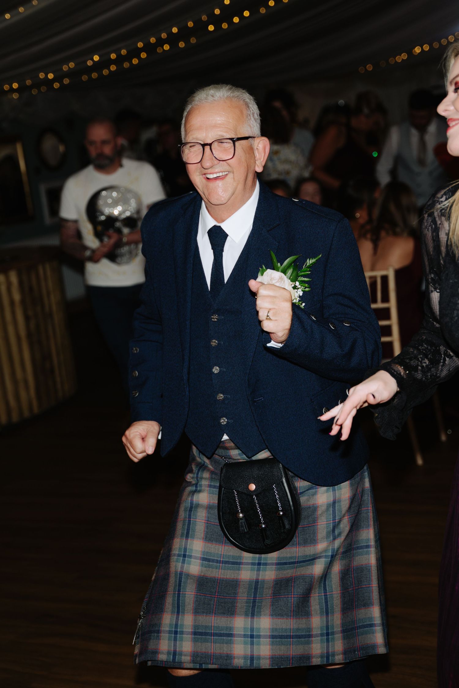 Man in traditional Scottish kilt and navy jacket dancing joyfully at wedding reception with string lights overhead.