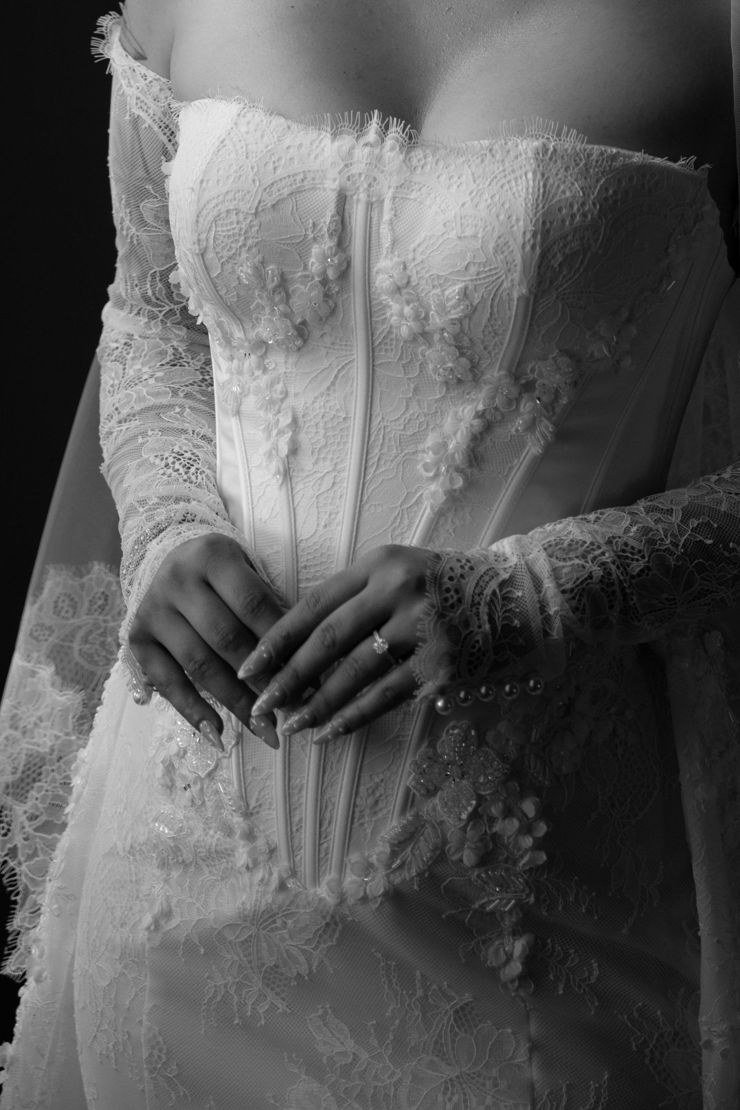 Black and white photo of a bride in an elegant lace corset wedding gown with floral details, hands folded gracefully.