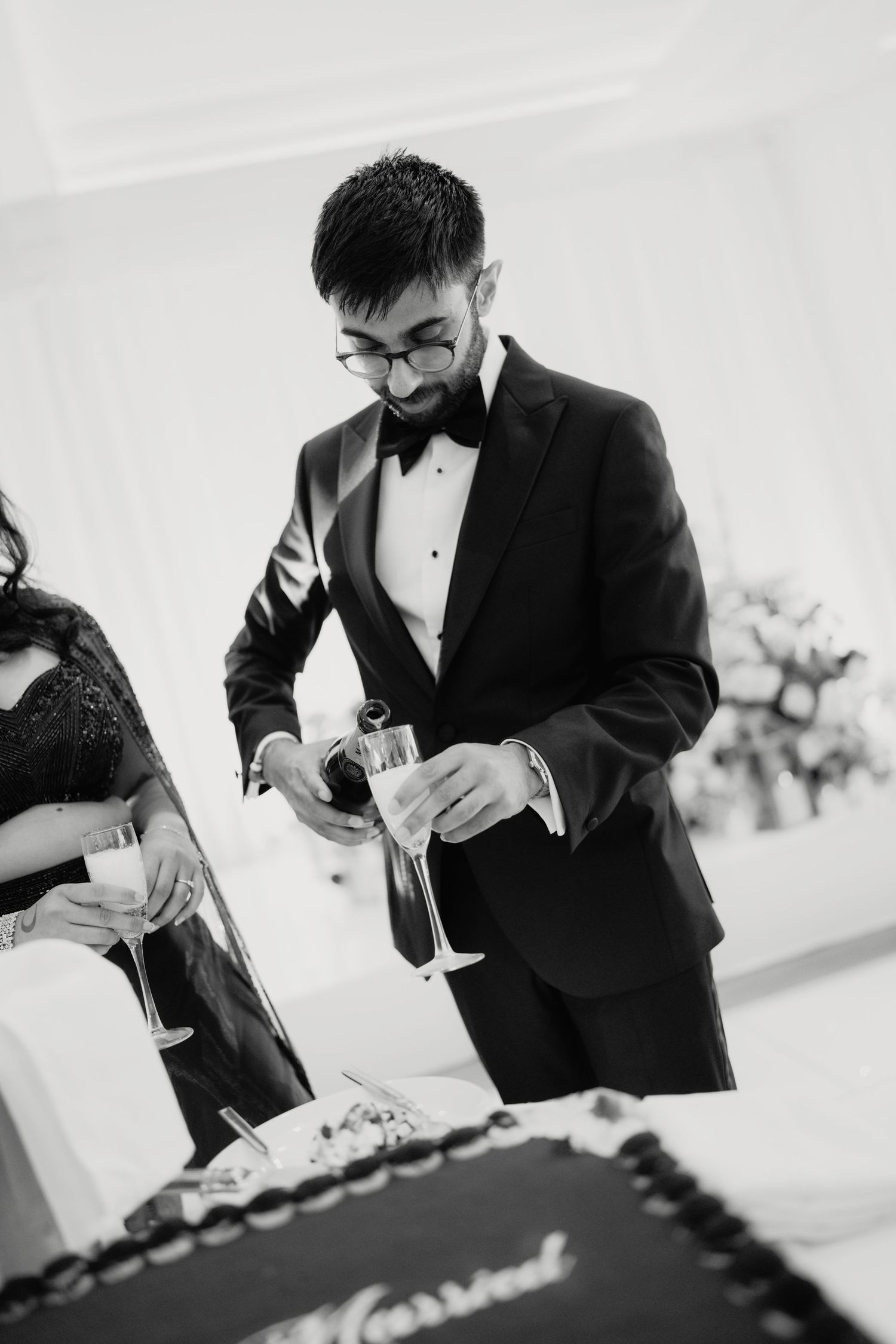 Groom in tuxedo pouring champagne at wedding reception cake table, black and white photo.