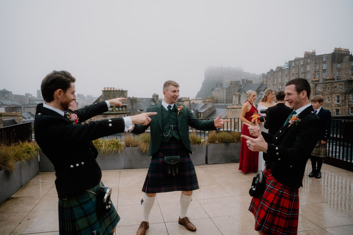 Wedding party wearing kilts dance together on a rooftop overlooking Edinburgh's historic architecture.