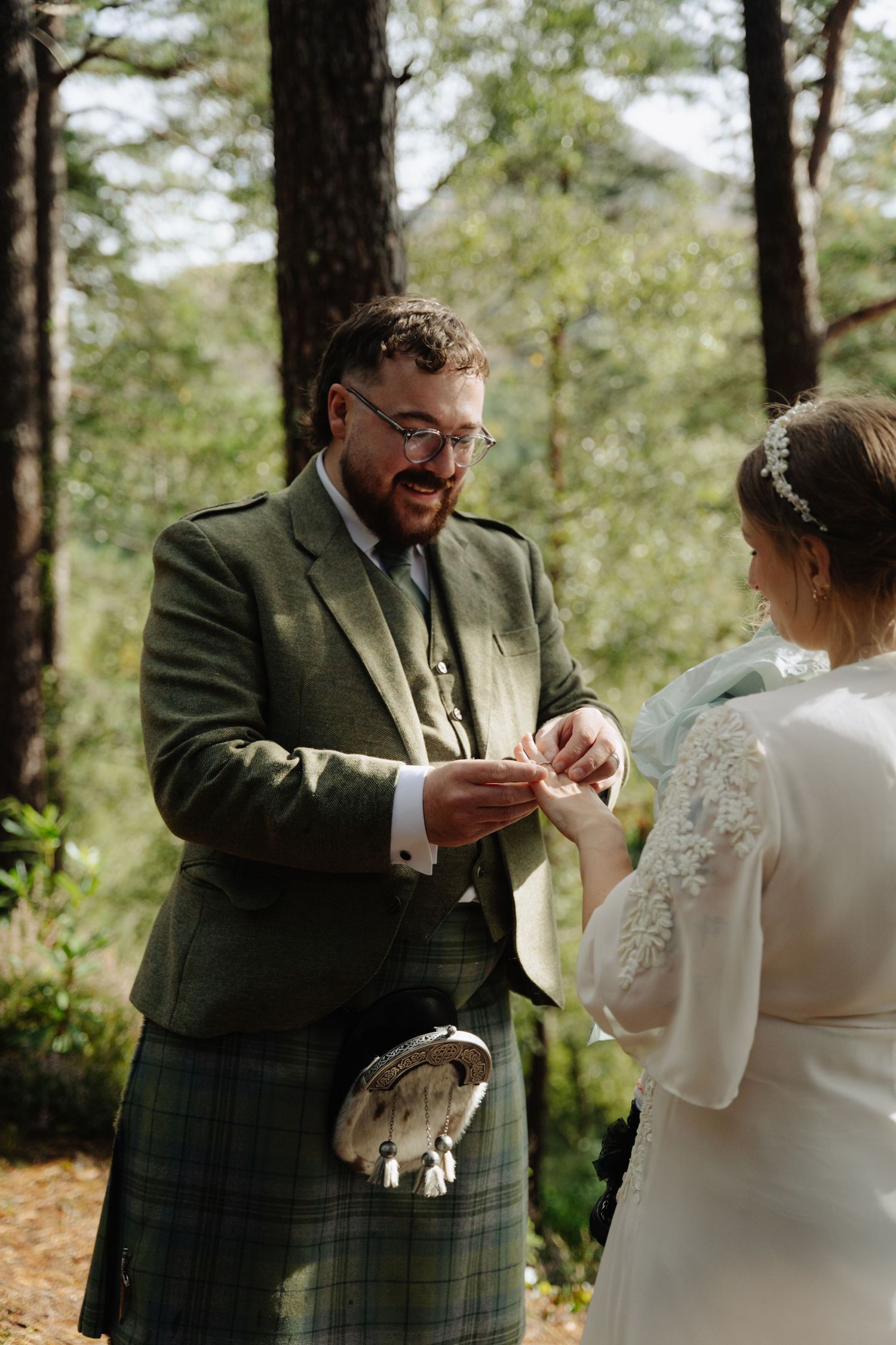 A couple exchanges rings during an outdoor wedding ceremony in a wooded forest setting.