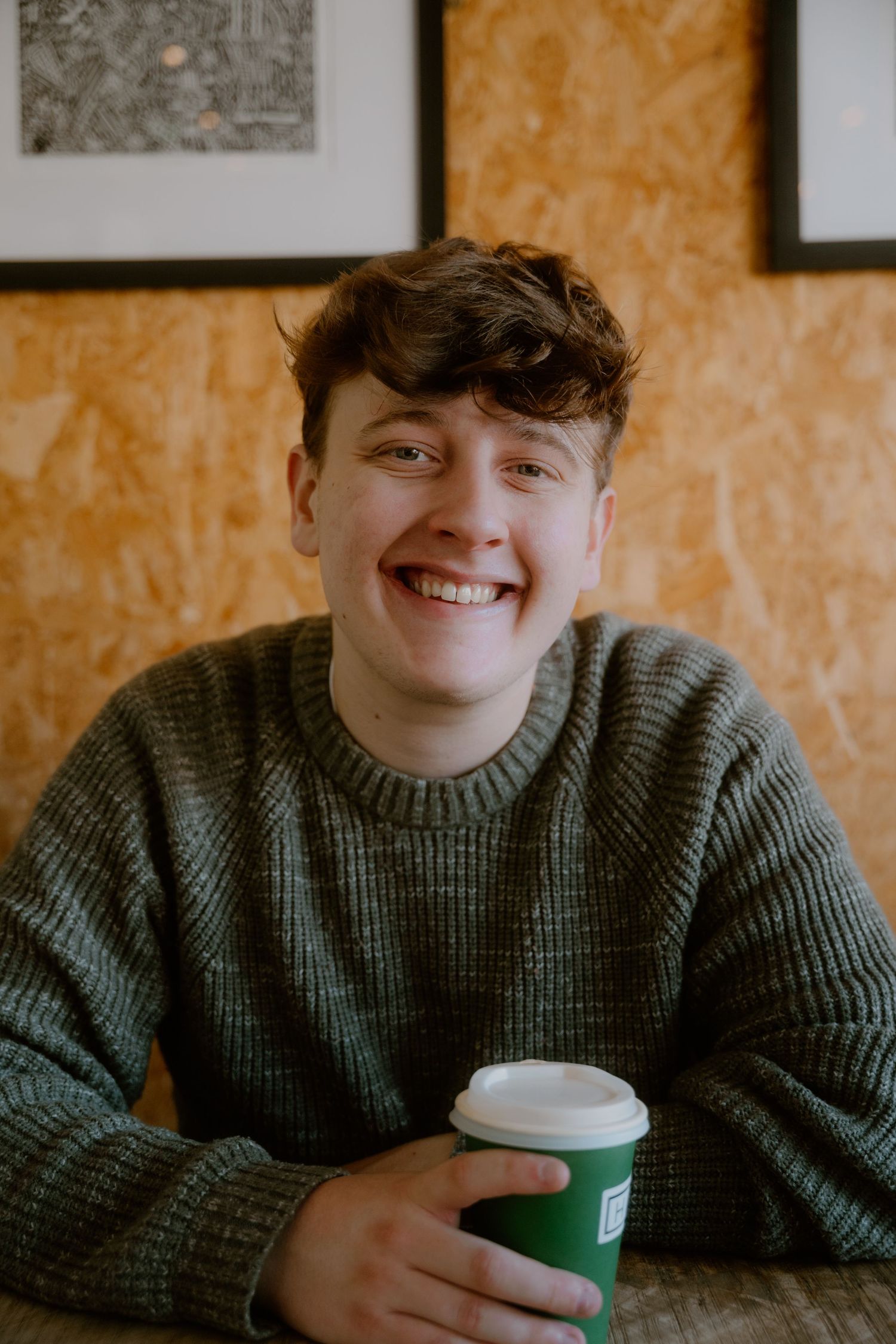 Someone wearing a cozy green sweater smiles while holding a takeout coffee cup against an orange textured wall.