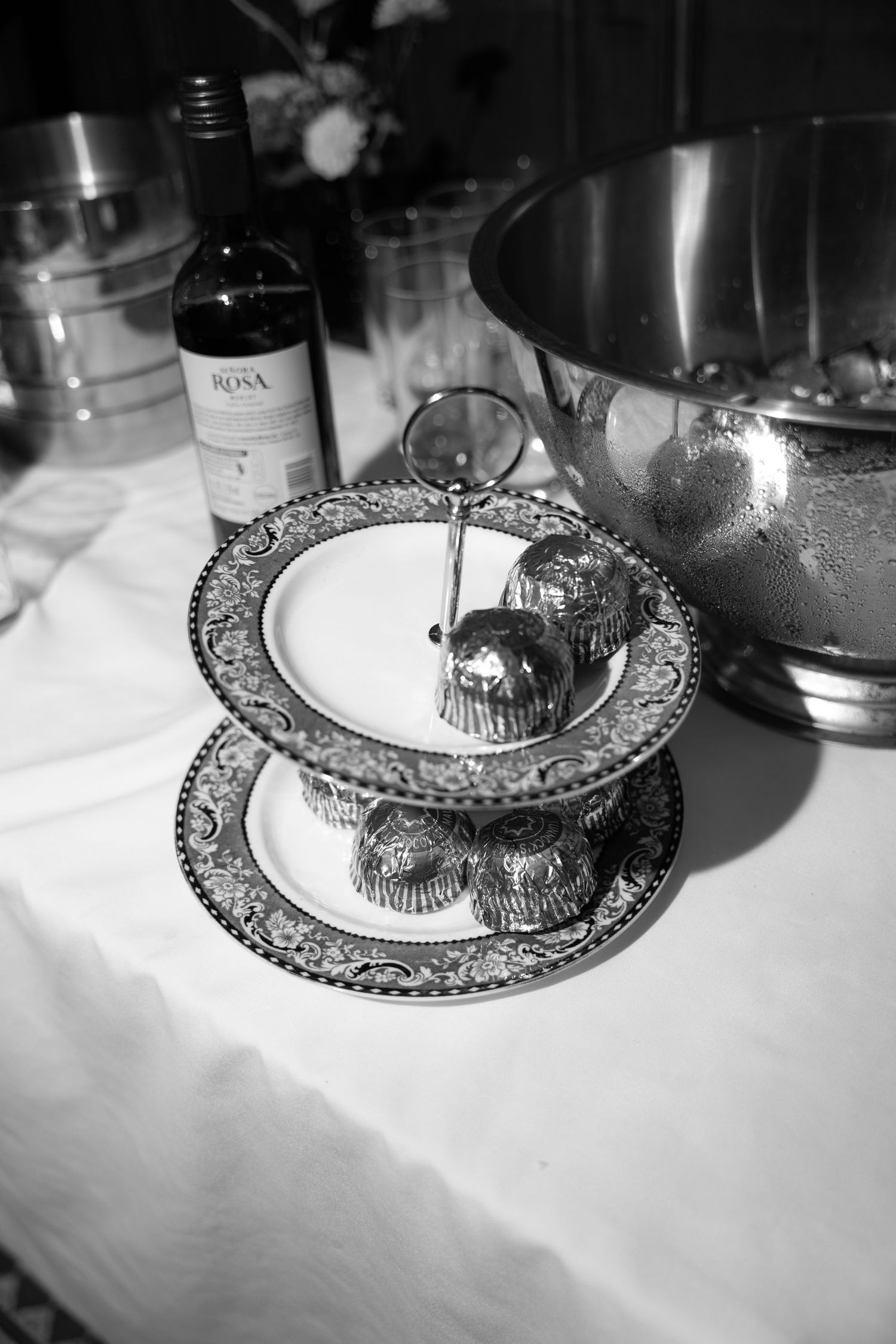 Black and white photo of elegant two-tier cake stand with cupcakes, wine bottle, and silver ice bucket on white tablecloth.
