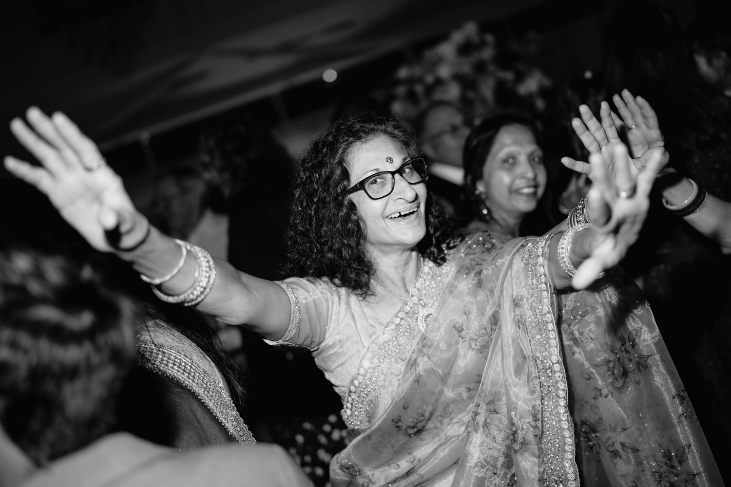 Woman in saree dancing joyfully with arms raised at an Indian wedding celebration, black and white photo.