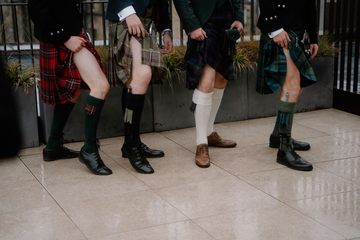 Four pairs of legs wearing kilts and boots show traditional Scottish attire at a formal gathering.