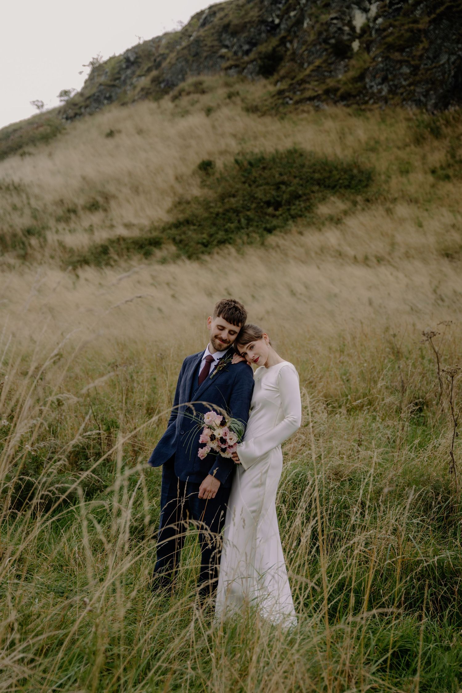 Bride and groom embracing in tall grass field with rocky hillside backdrop on their wedding day.