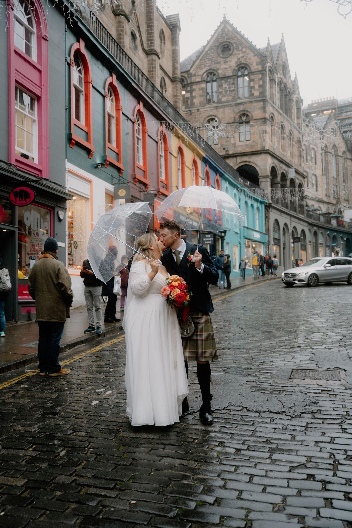 A couple shares a romantic moment under an umbrella on a rainy cobblestone street in Edinburgh.