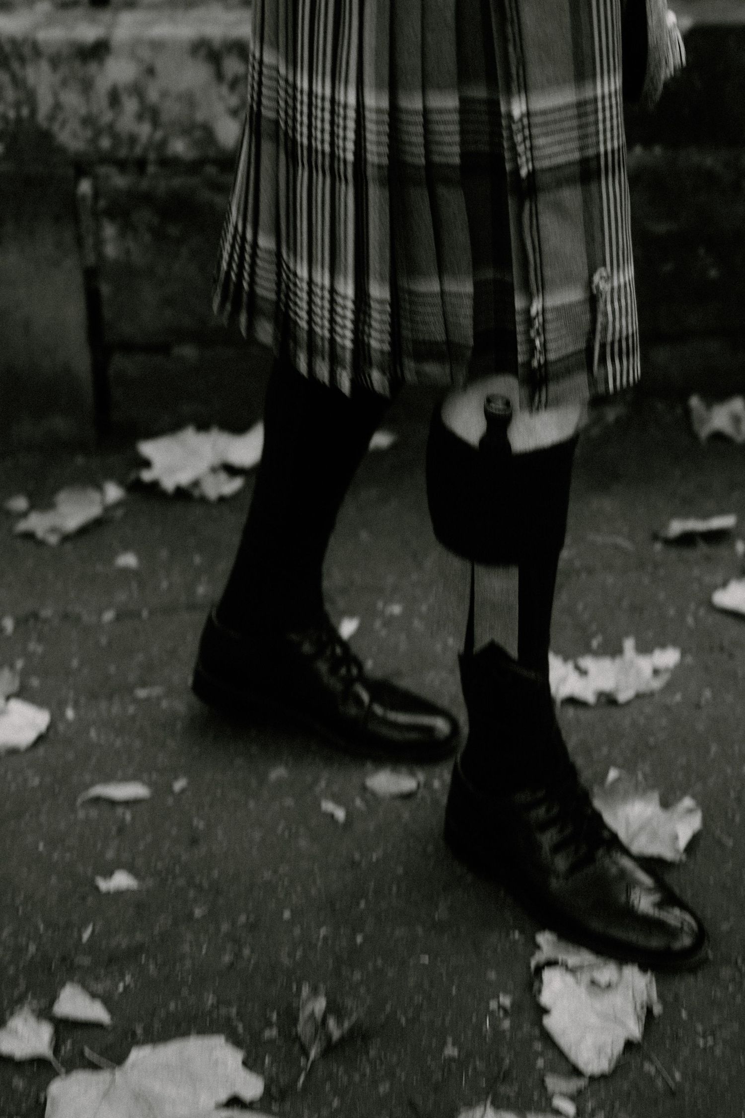 Black shoes and a traditional plaid kilt hem visible against fallen leaves on pavement.