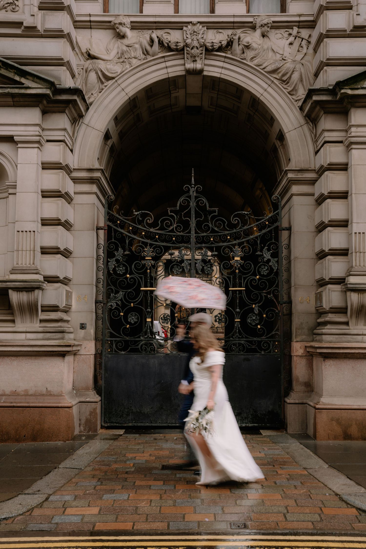Blurred bride and groom walking past ornate iron gates of a grand stone building on a rainy wedding day.