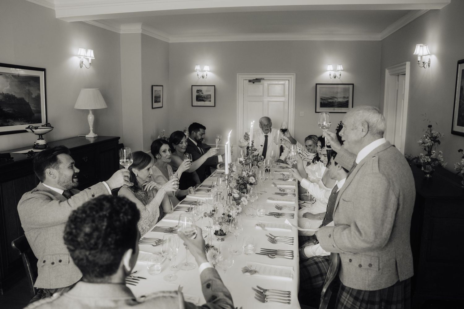 Black and white photo of wedding guests raising glasses in a toast at a long formal dining table with candles.