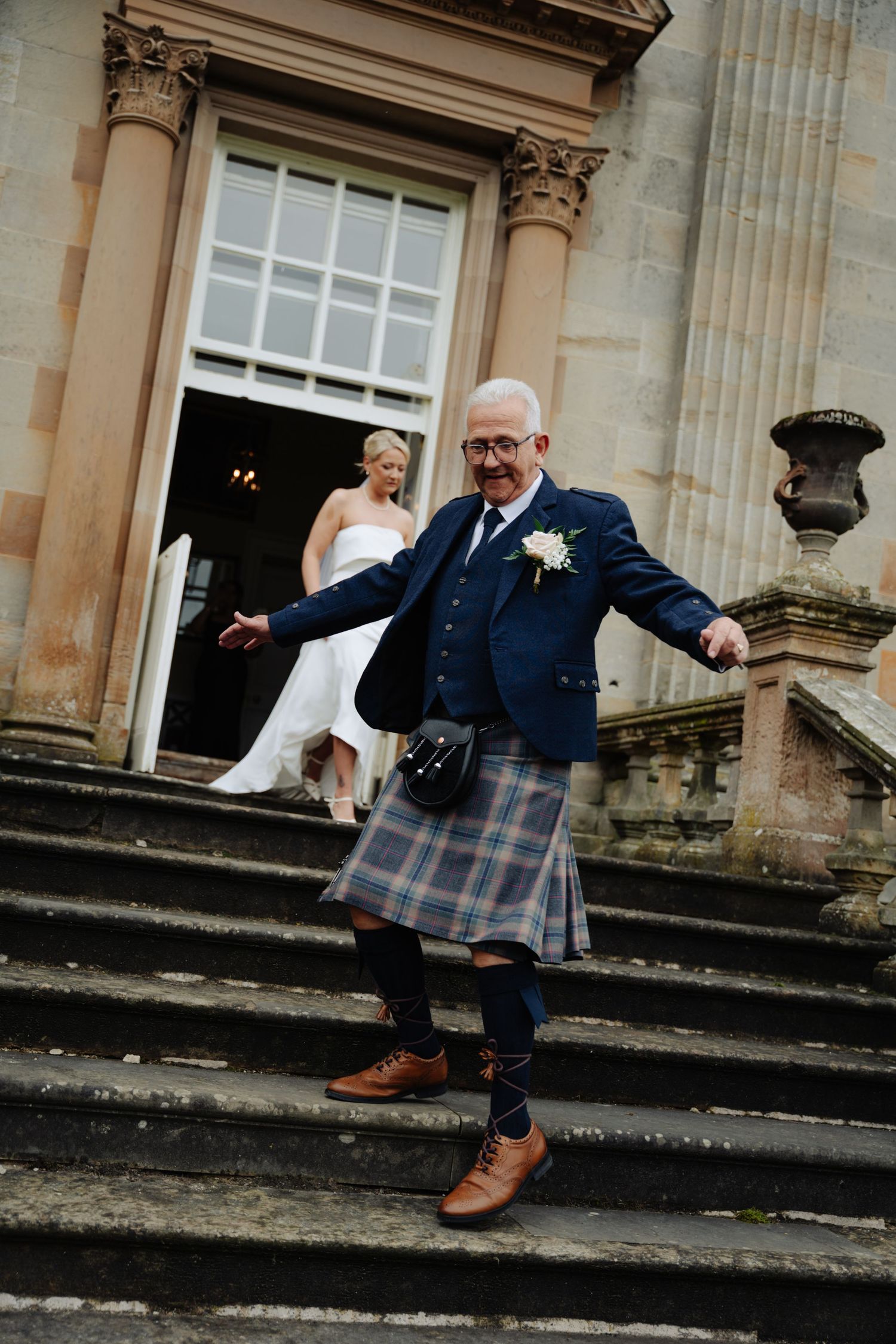 A groom in a navy suit and kilt playfully jumps down stone steps at a historic building entrance. Boswell Estate