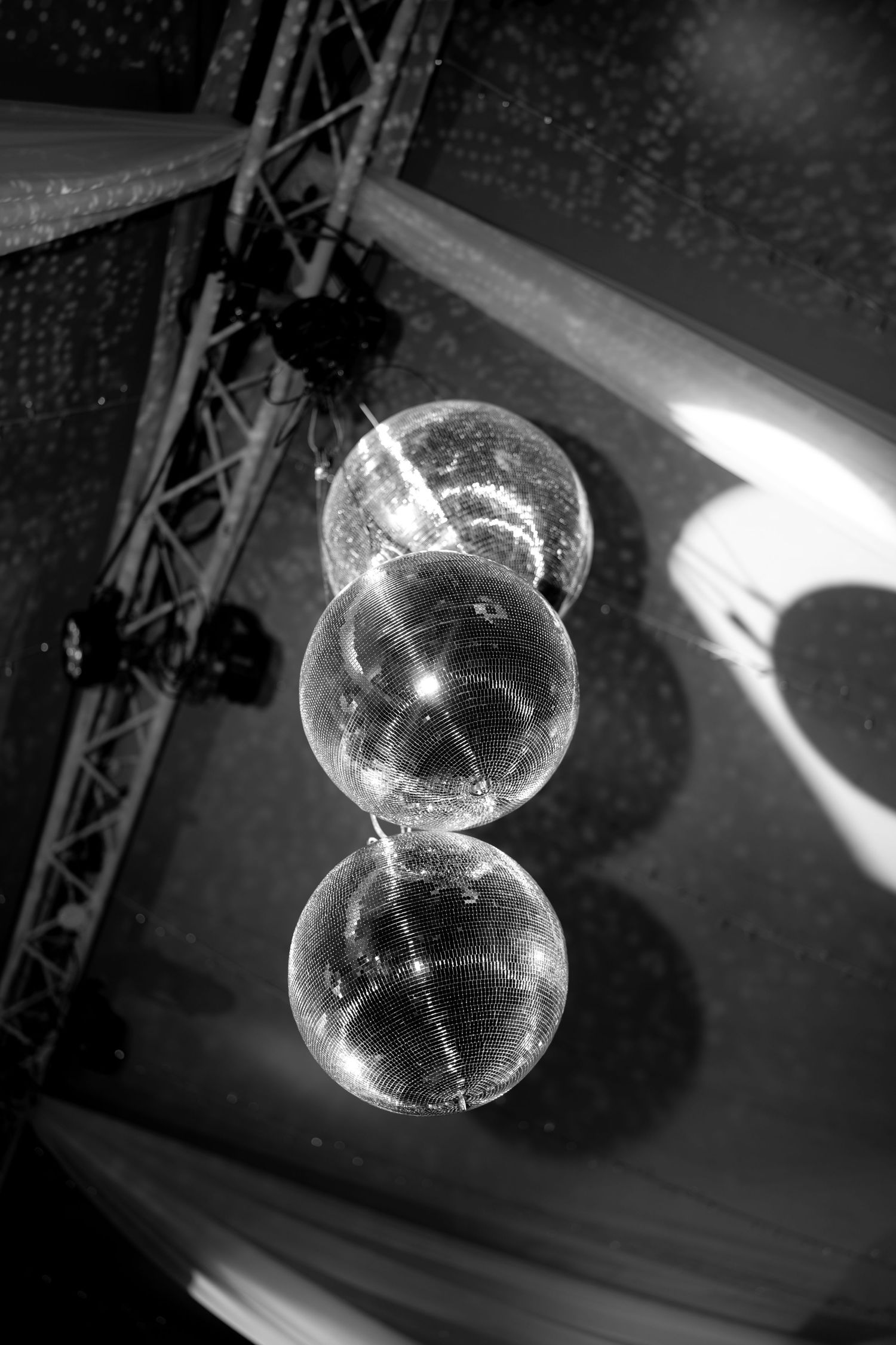Black and white photo of three stacked disco mirror balls hanging from a metal truss ceiling at an event venue.