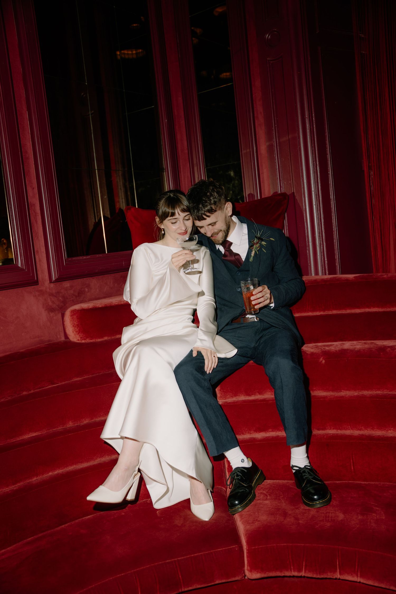 Bride and groom sit intimately on red velvet curved sofa, sharing drinks in elegant dark venue.