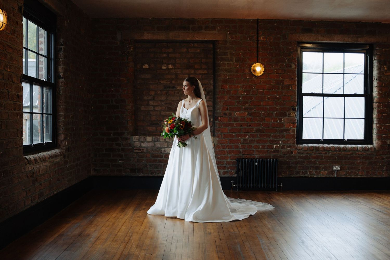 A bride in a flowing white gown stands gracefully in a rustic brick loft space with natural window lighting. Glasgow Engine Works
