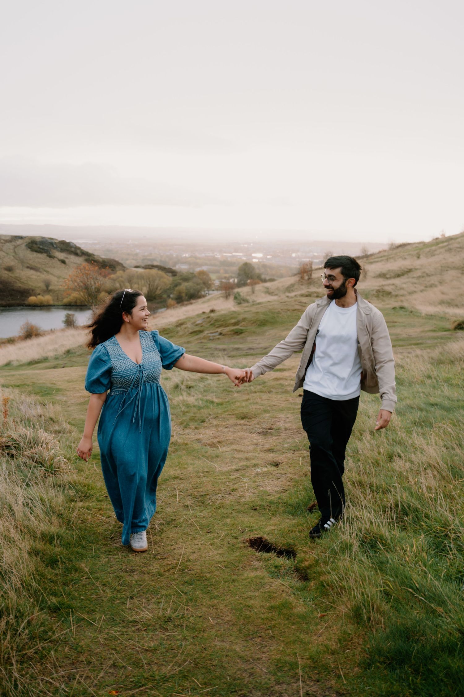A couple holds hands while walking through a scenic grassy hillside on an overcast day.