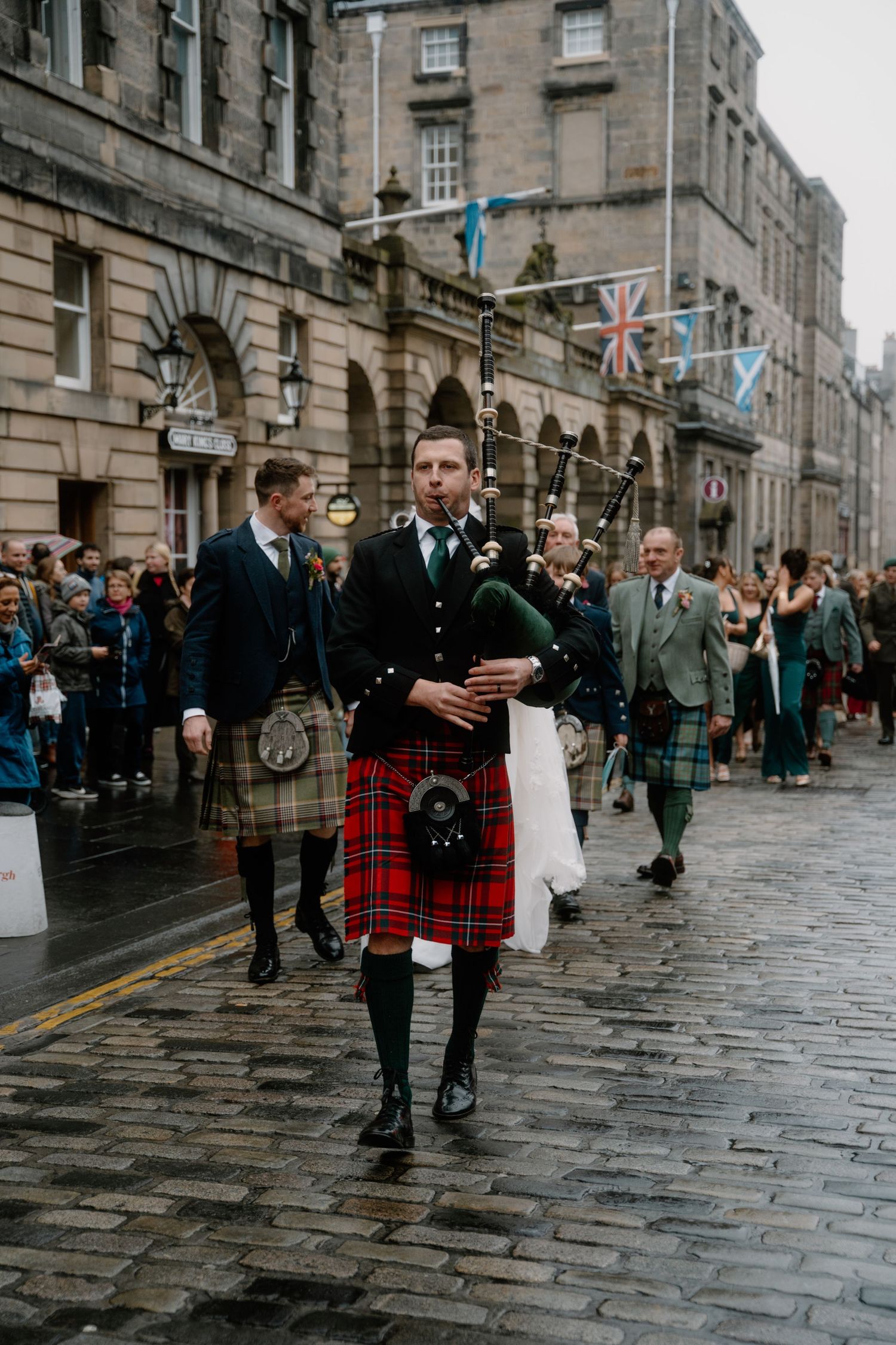 Kilted musicians walk along historic Royal Mile with Scottish flags and stone architecture.