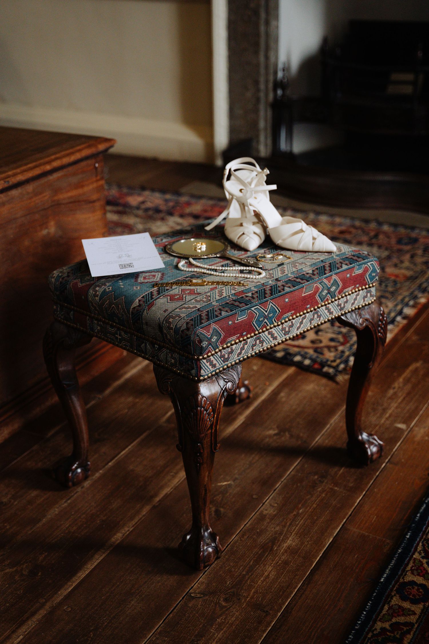 Vintage footstool with ornate tapestry upholstery and carved wooden legs sits on hardwood floor in moody lighting. Boswell Estate