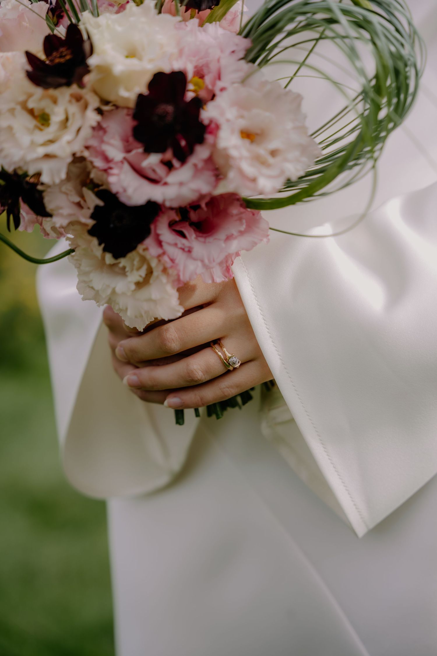 Bride holding a pink and burgundy wedding bouquet with green accents, wearing a gold ring on her finger.