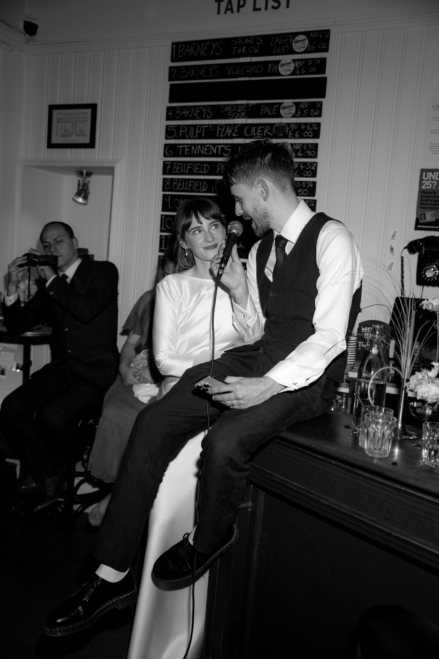 Black and white photo of a couple sharing a romantic moment at a bar, with a tap list chalkboard visible behind them.