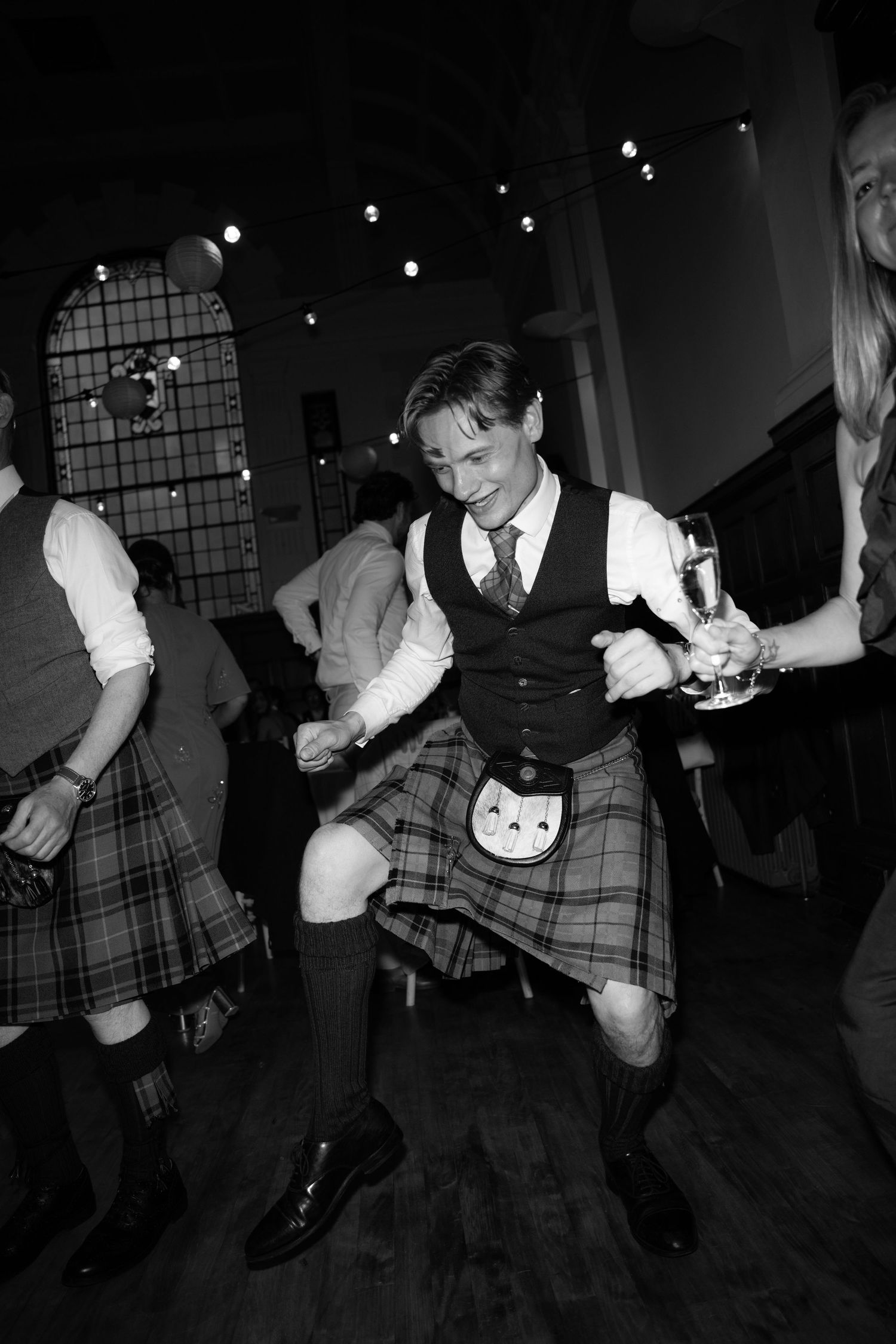 Man in traditional Scottish kilt dancing joyfully at a celebration in black and white photography.