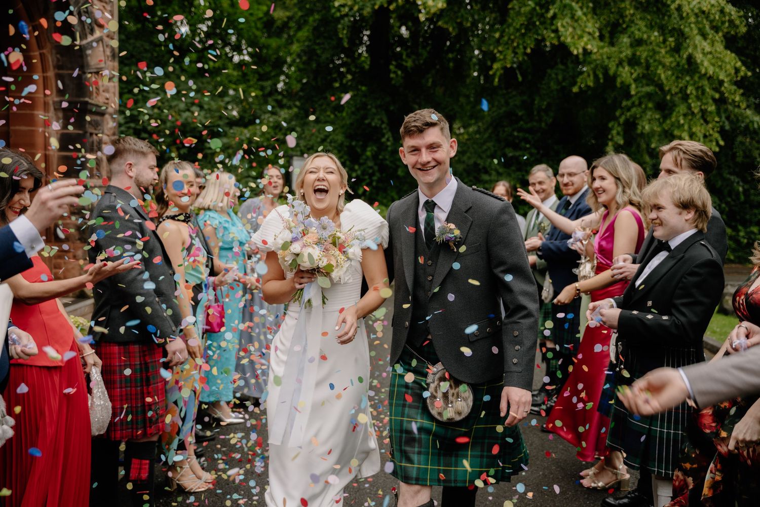 Bride and groom walk through colorful confetti shower with smiling wedding guests lining the path outdoors.