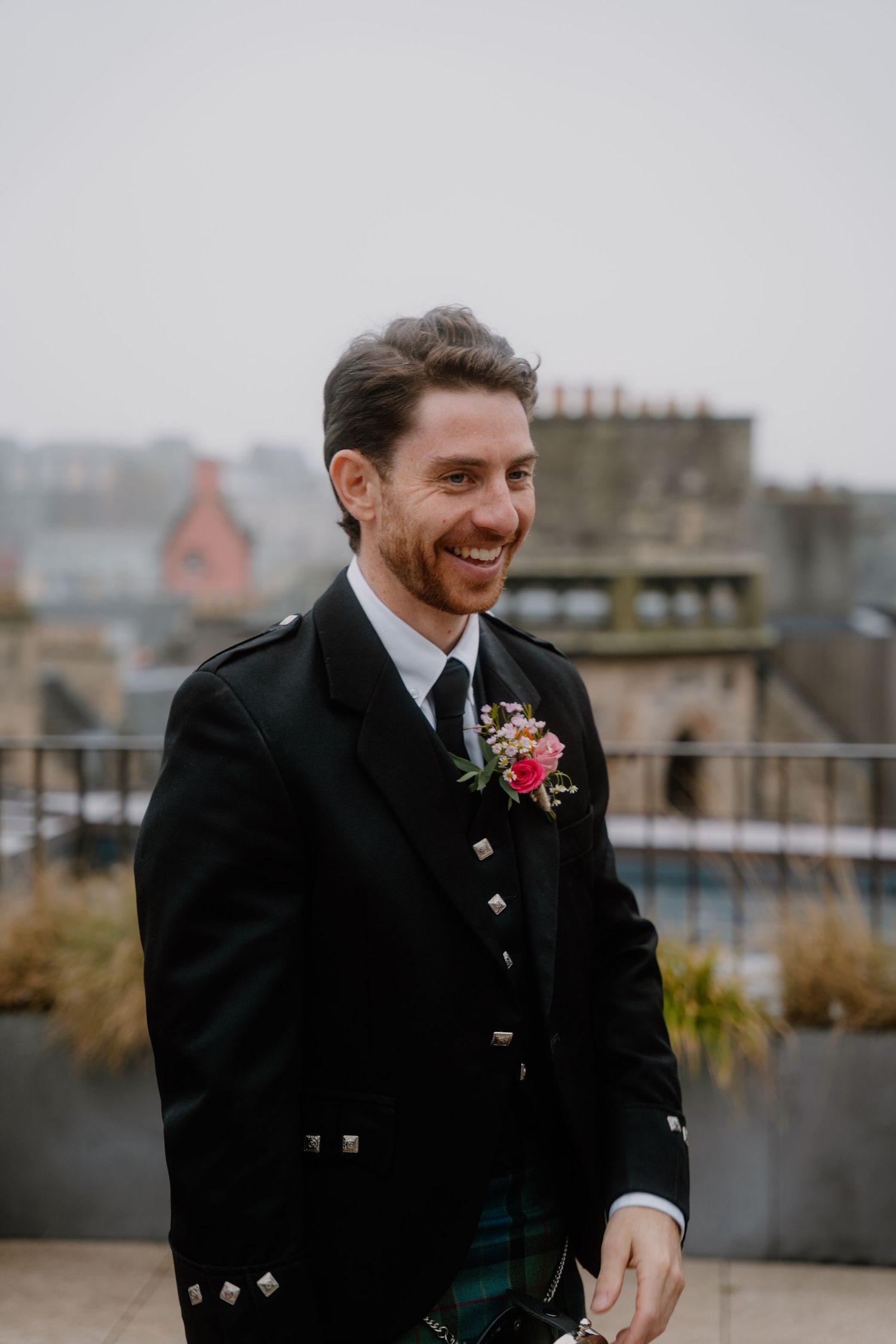 A person wearing formal Scottish attire with a boutonniere smiles on a rooftop terrace.