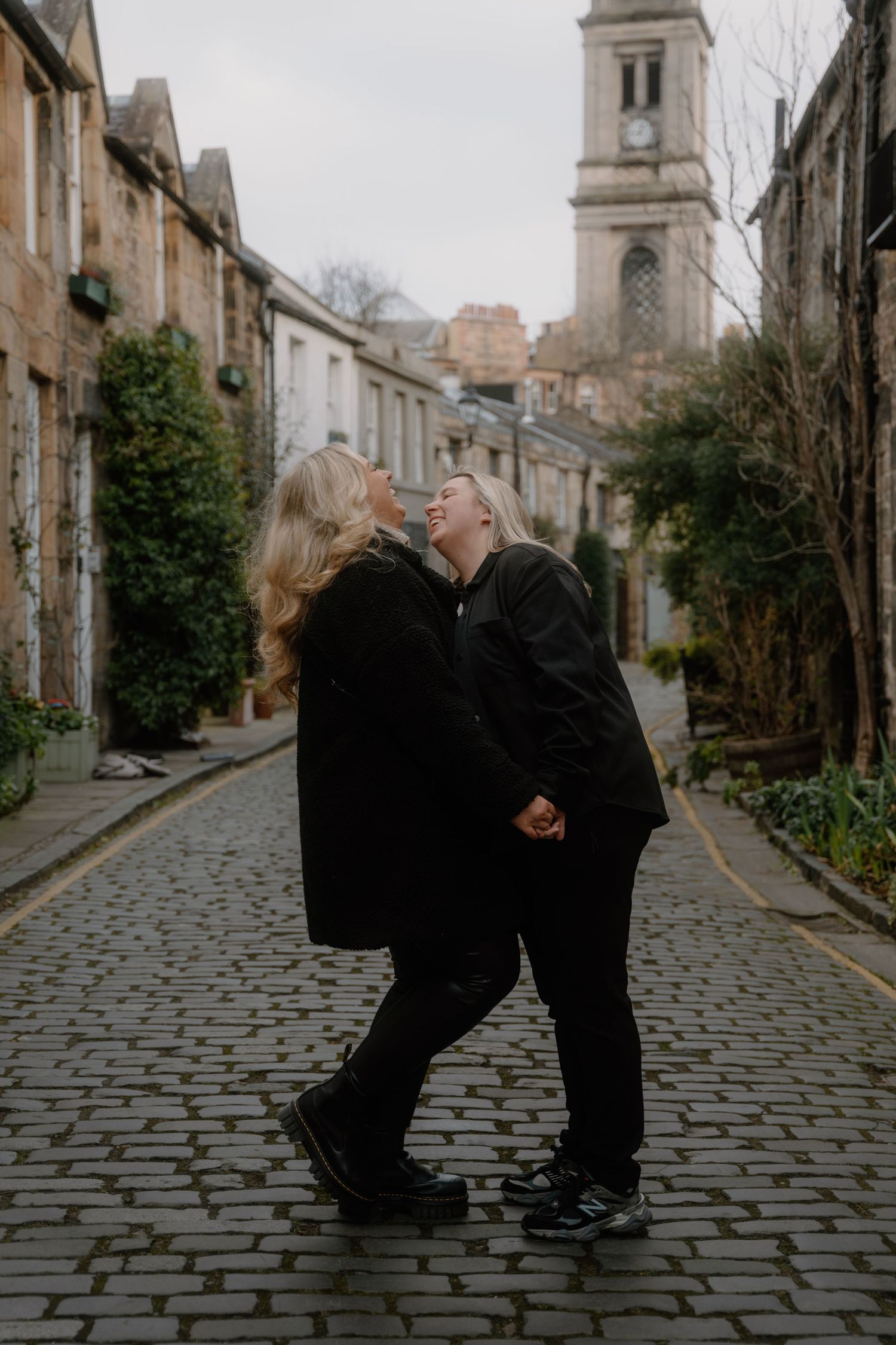 A romantic couple in black coats sharing an intimate moment on a cobblestone street with a church tower in the background.
