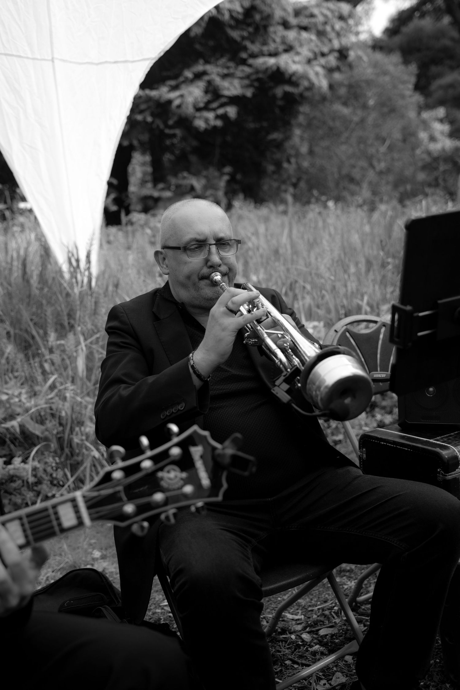 Bald man in black suit playing trumpet outdoors in black and white photo with guitar in foreground.