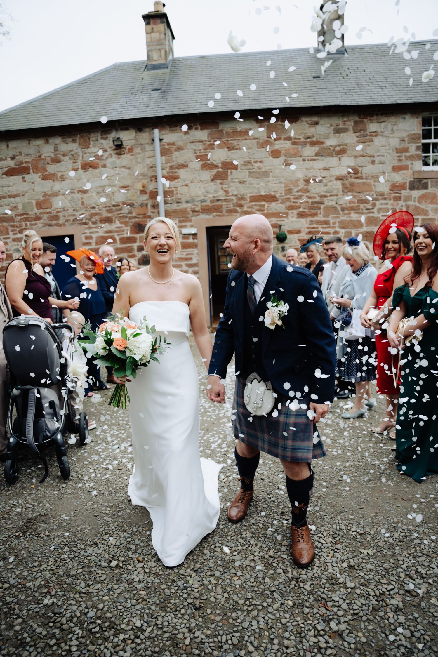 A wedding celebration at a rustic stone building with confetti being thrown as a couple in traditional Scottish attire walks. Boswell Estate