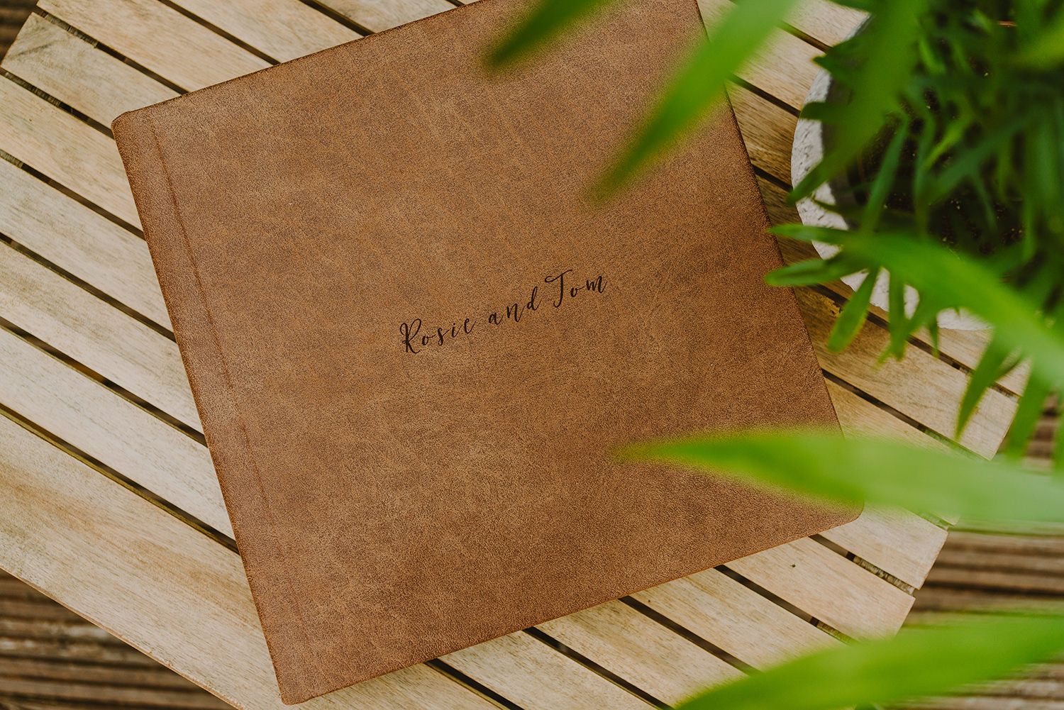 Brown leather photo album on wooden slats with green plant leaves in frame.