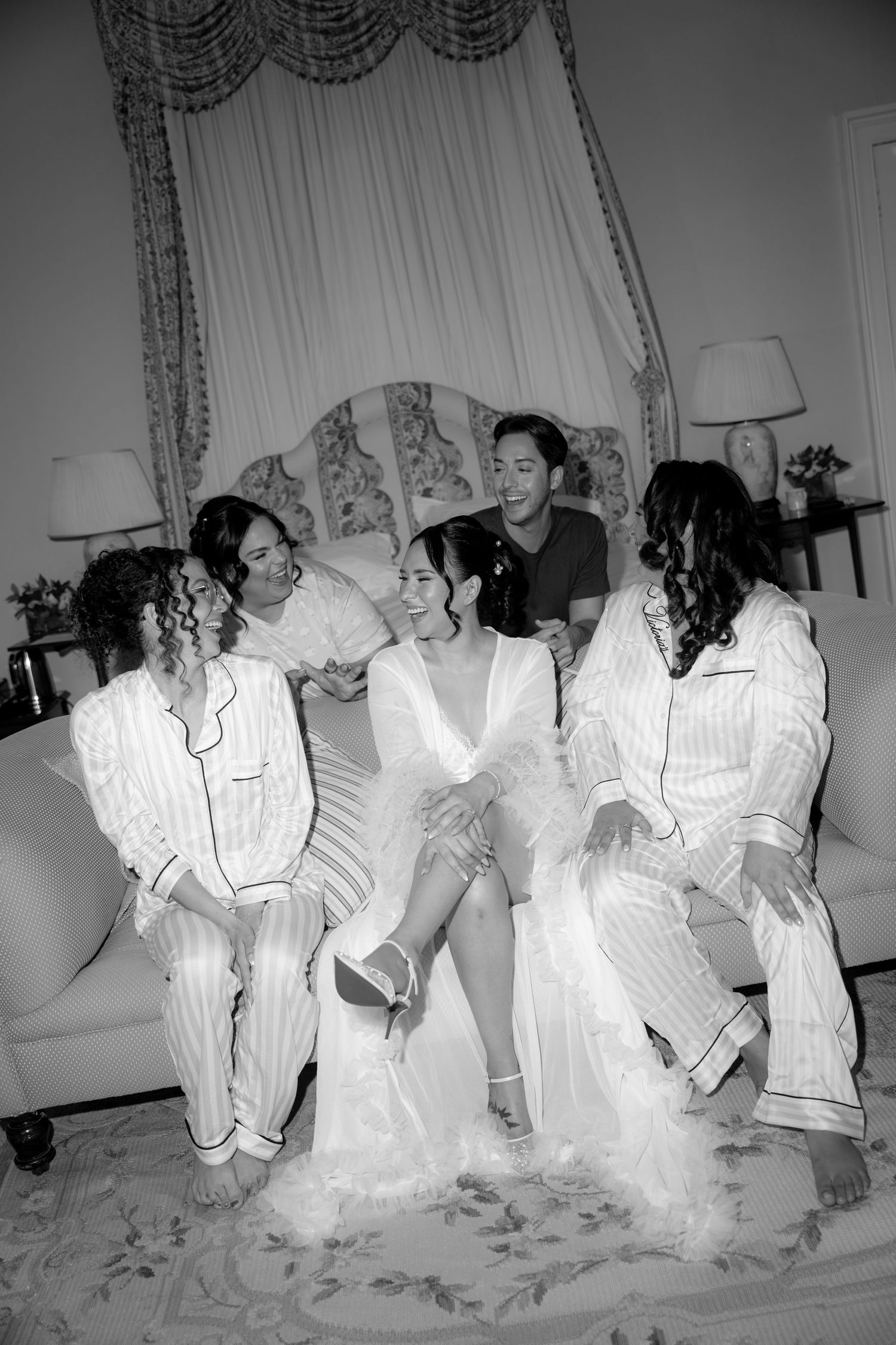 Black and white photo of a bride in white robe with bridesmaids in matching pajamas laughing together on a sofa.
