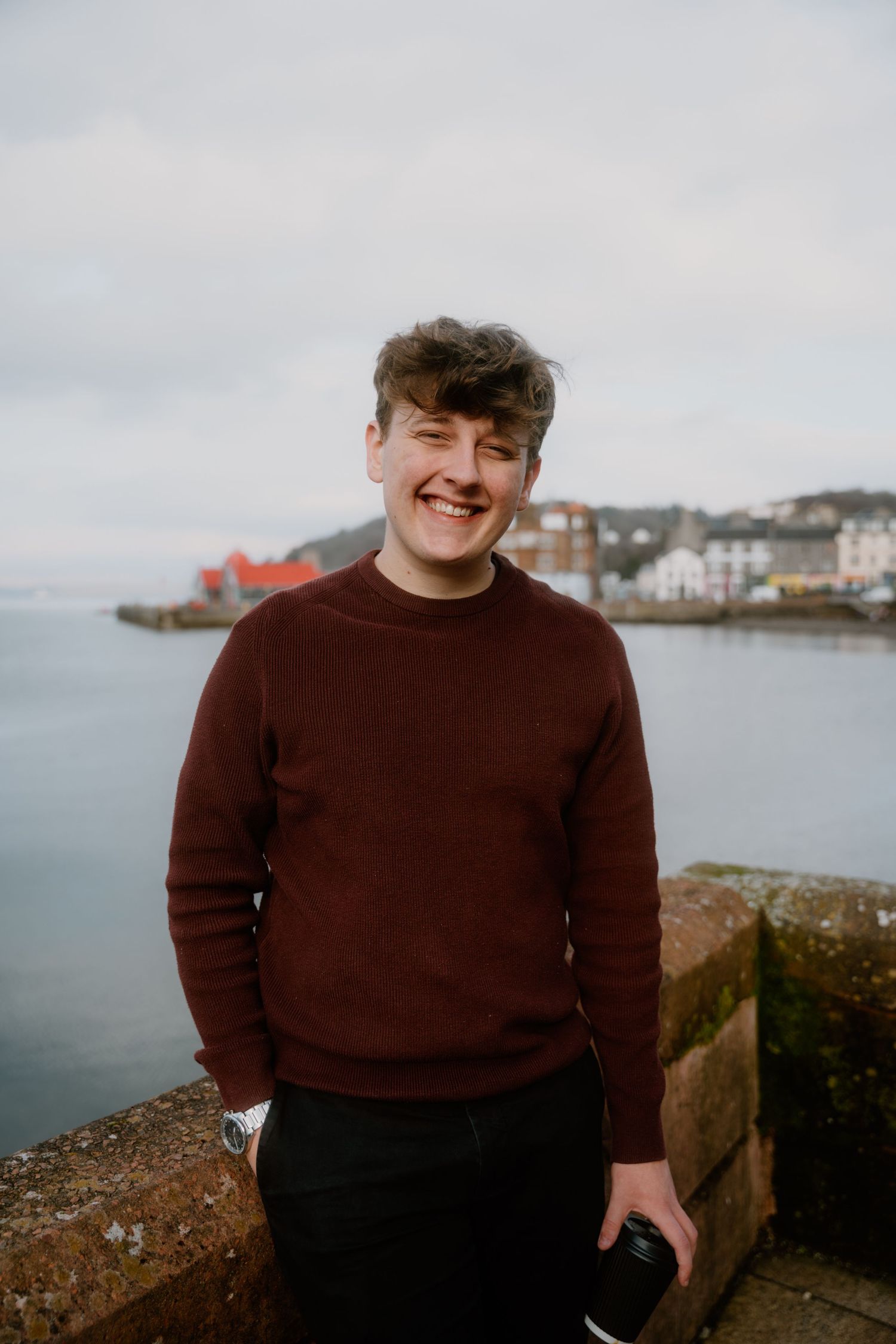 Person in brown sweater standing by coastal view with houses in background.