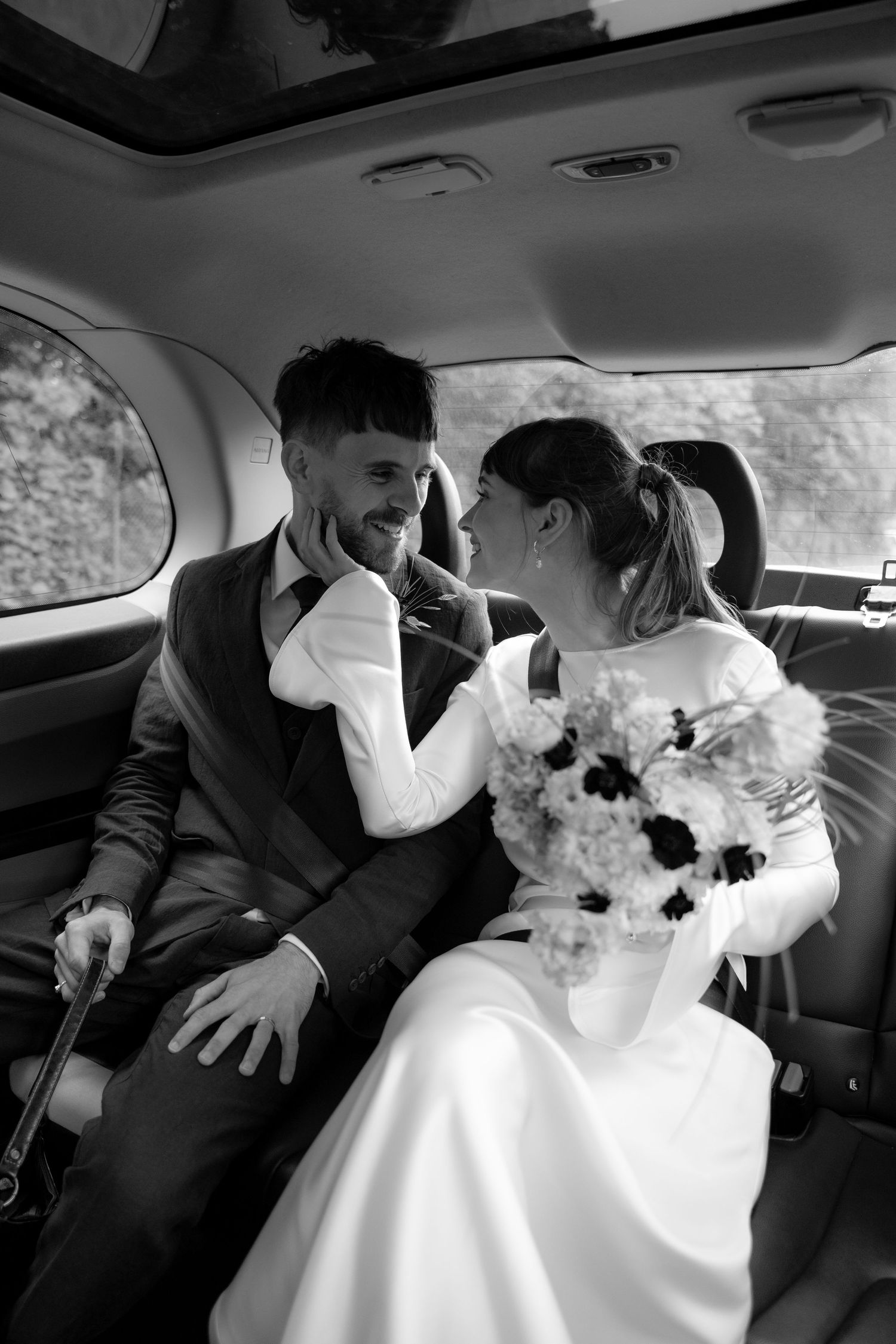Black and white photo of a smiling bride and groom sitting in a car backseat, bride holding a bouquet.