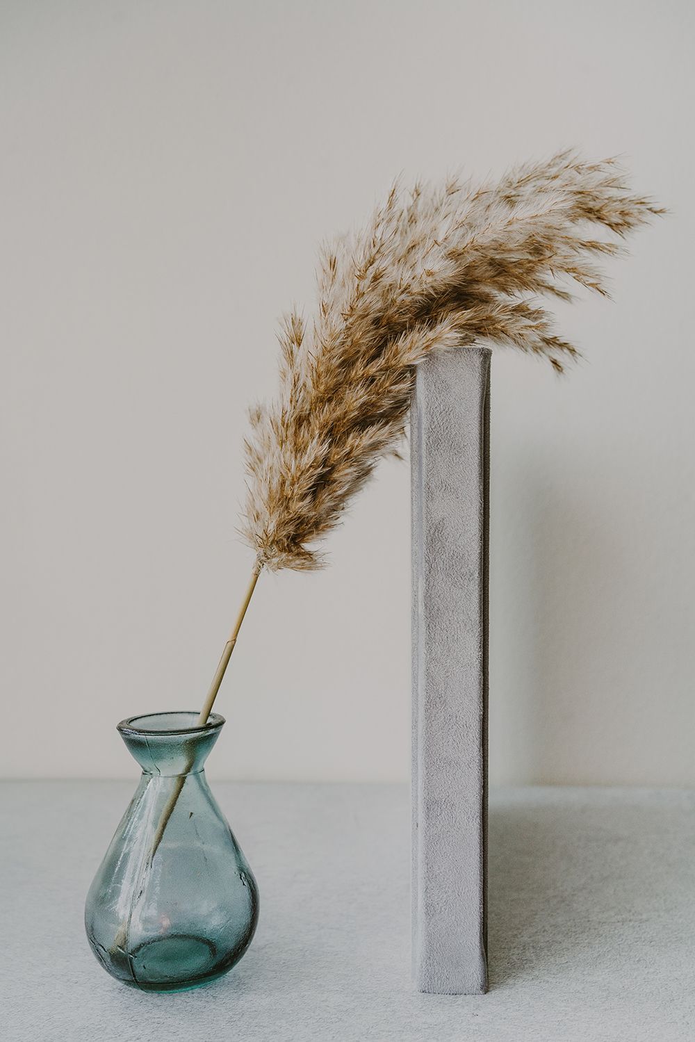 Dried pampas grass in a blue glass vase next to a white book against a neutral background.