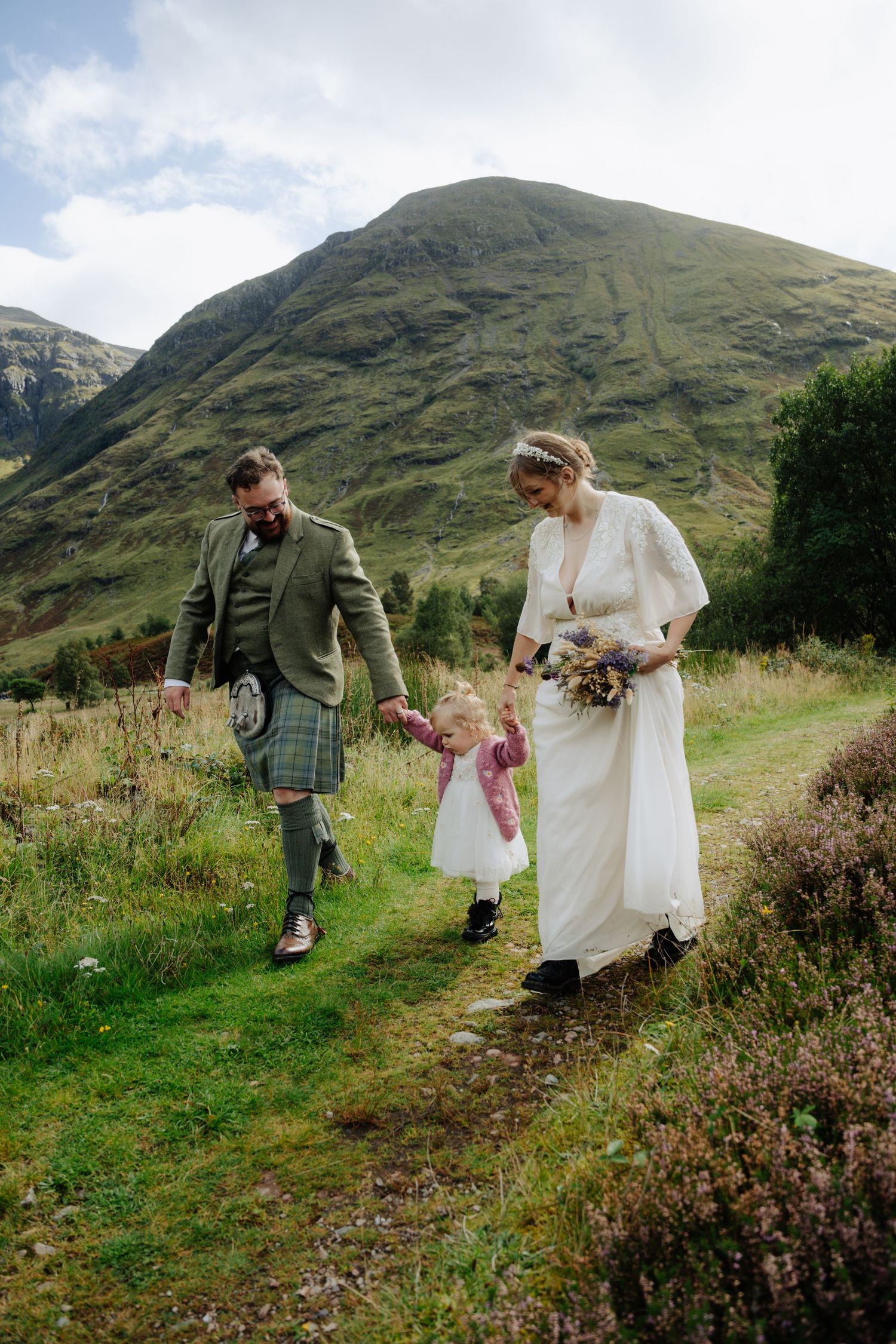 A couple walks through scenic Scottish highlands with mountains in the background during a wedding photoshoot.