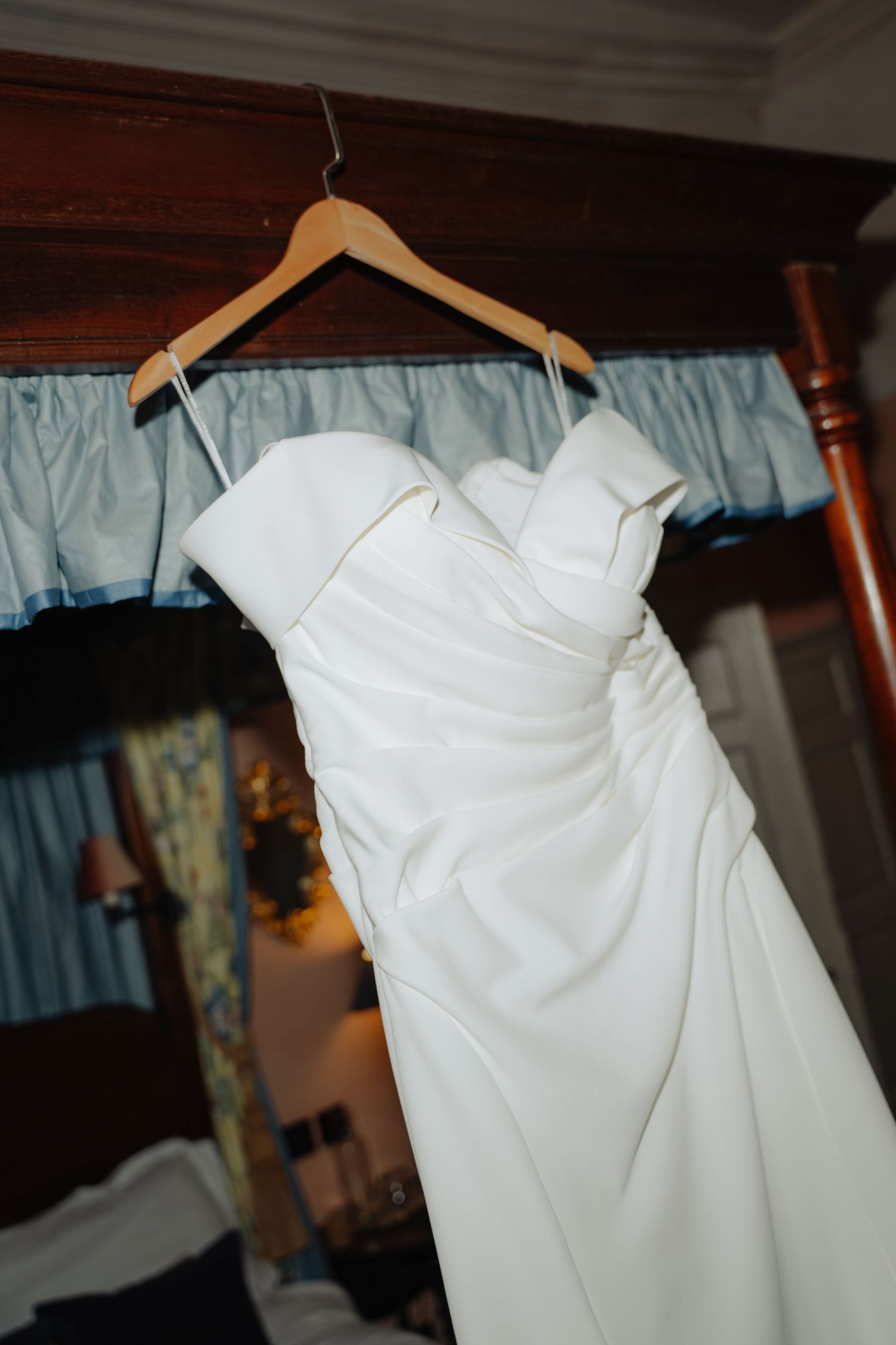 White strapless wedding dress hanging on wooden hanger in elegant bedroom with blue curtains.