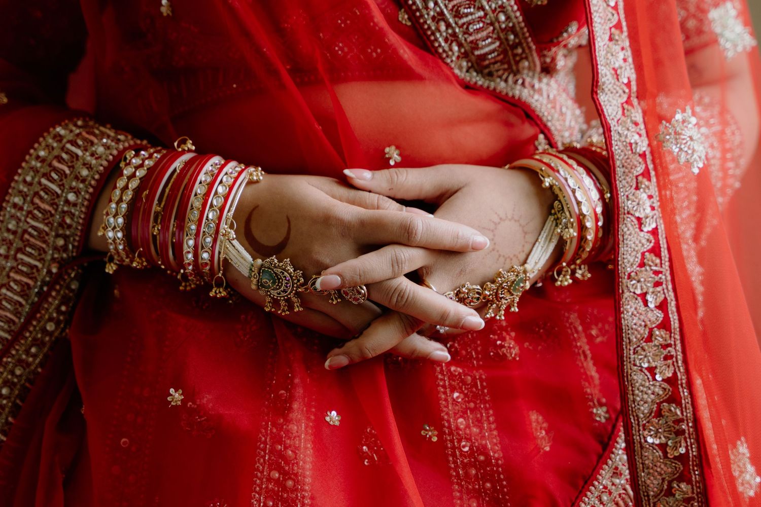 Close-up of Indian bride's hands adorned with gold bangles, red chura, and mehndi henna, wearing a red bridal lehenga.