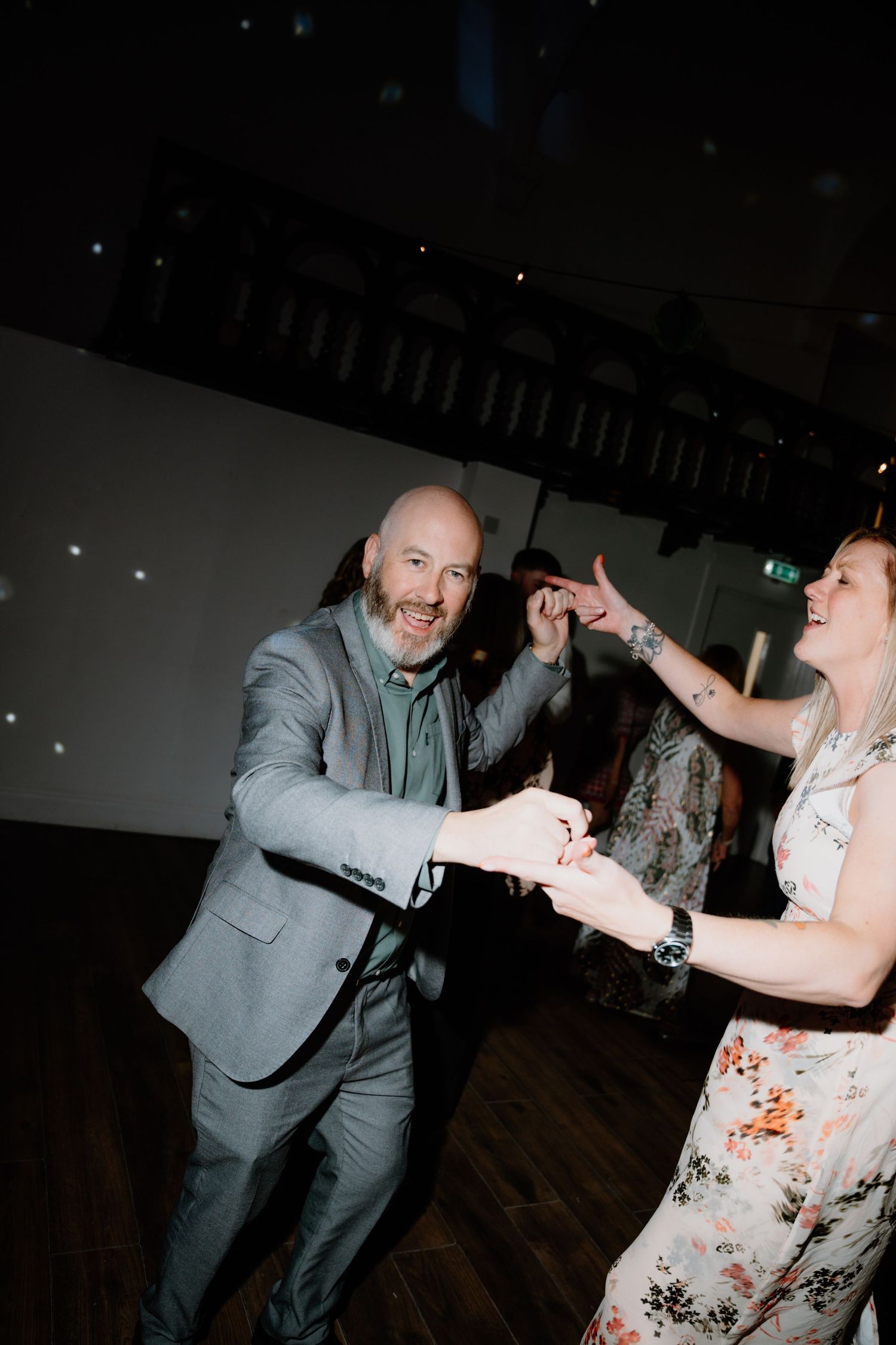 A couple dances together at a party, with the man wearing a gray suit and the woman in a floral dress.