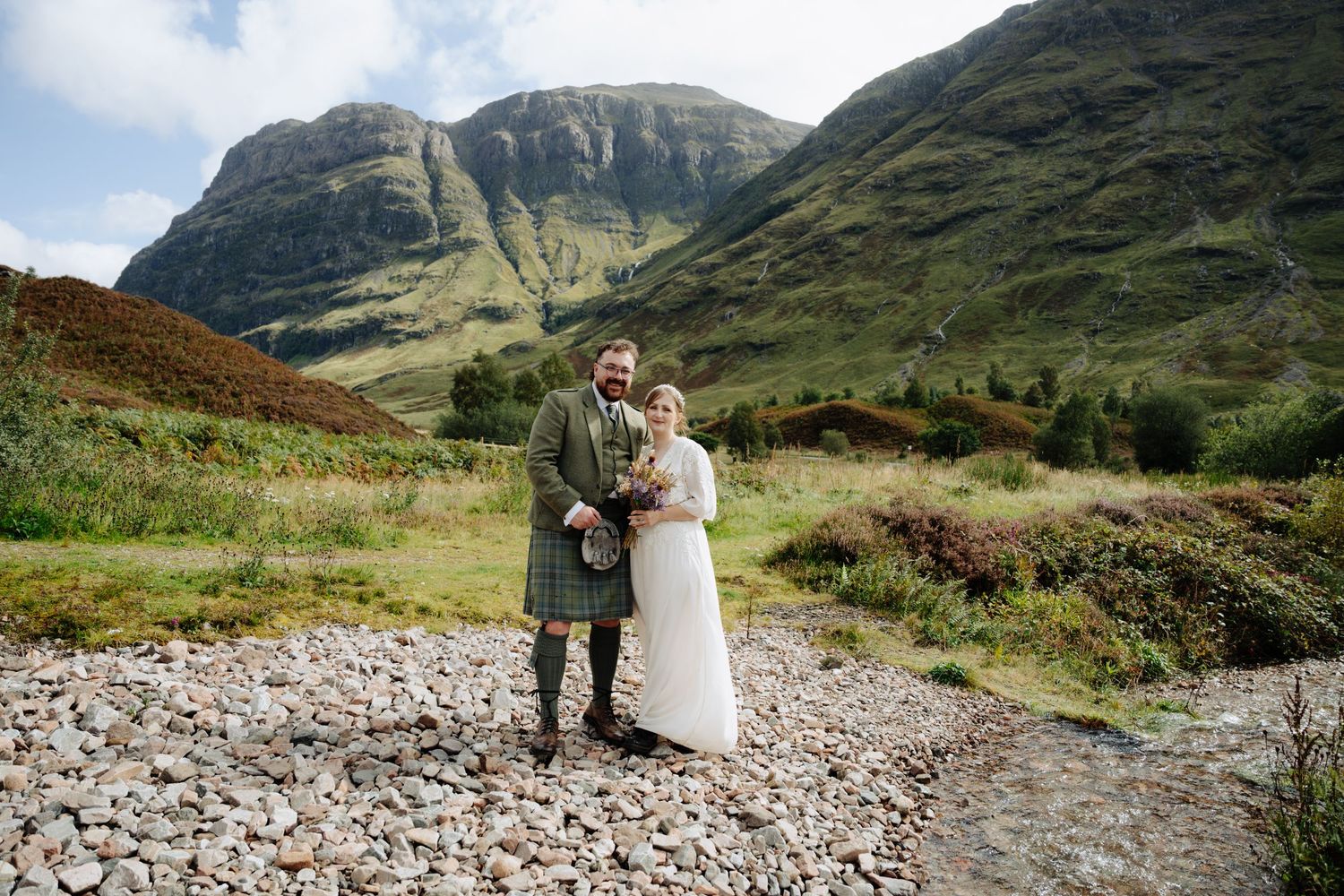 A couple stands together in the dramatic mountain landscape of Glen Coe, Scotland during their wedding photoshoot.