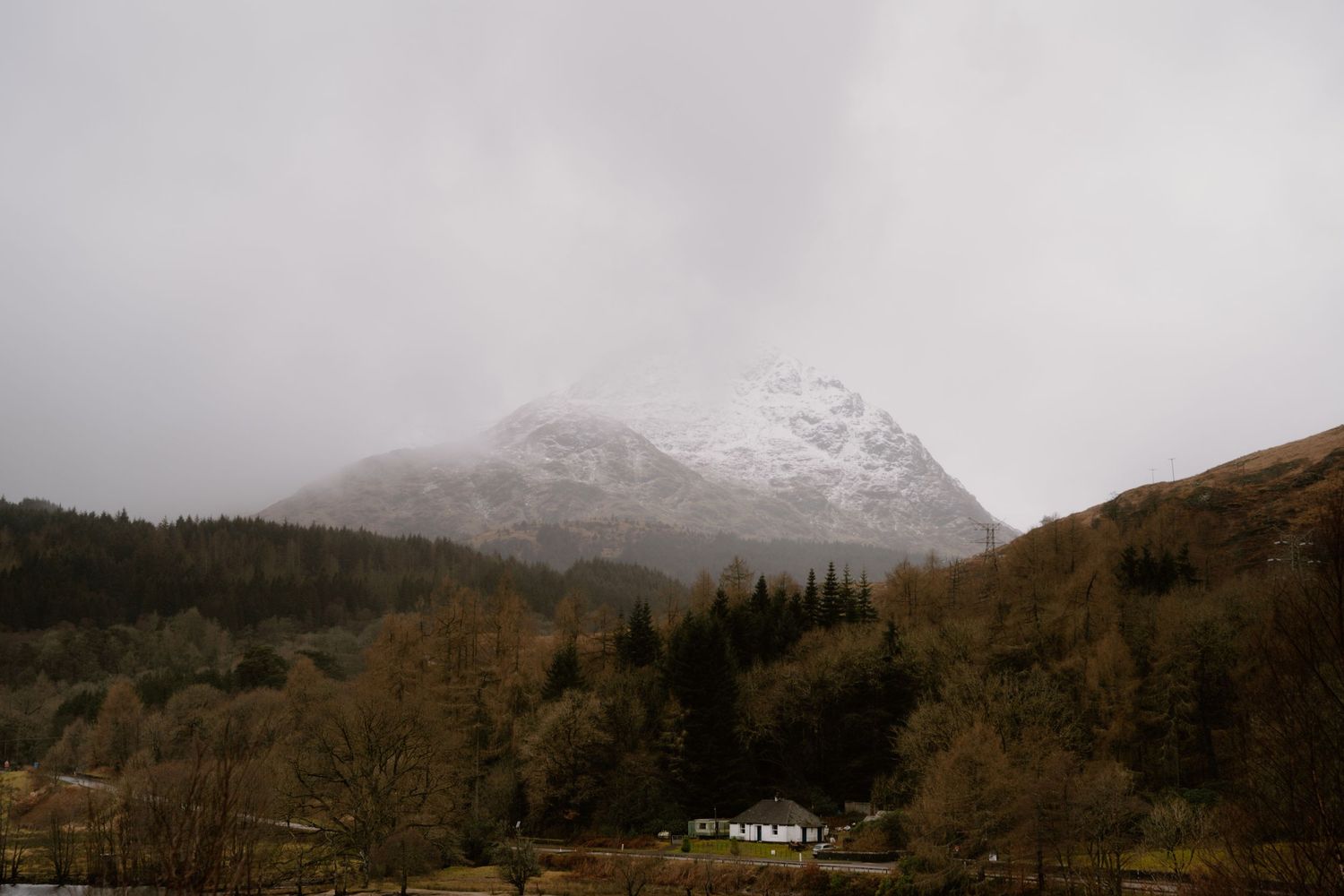 Snow-capped mountain peaks loom over a remote cottage nestled among autumn trees in misty Scottish highlands.