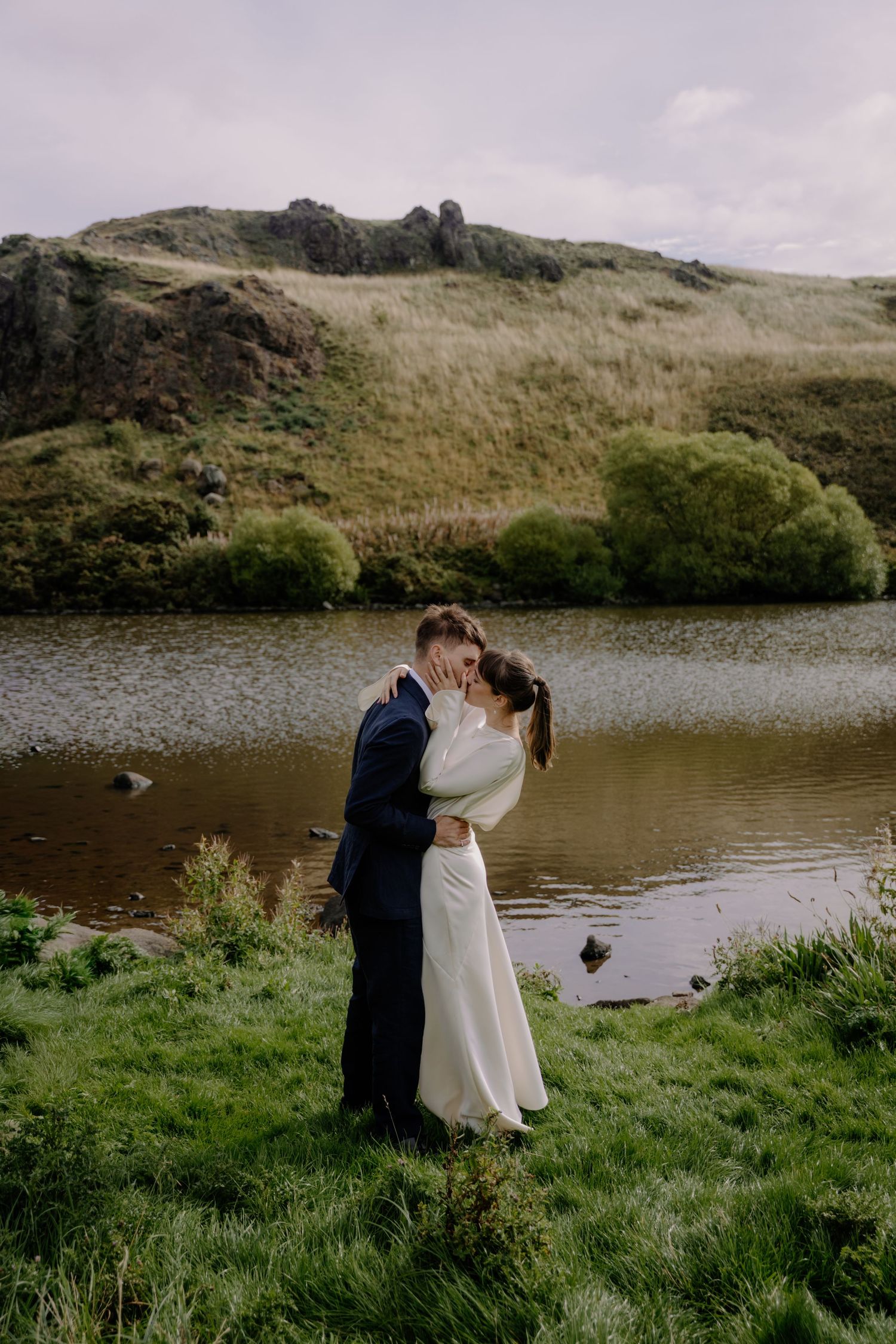 Couple sharing a romantic kiss in wedding attire beside a serene lake with lush green hills in the background.