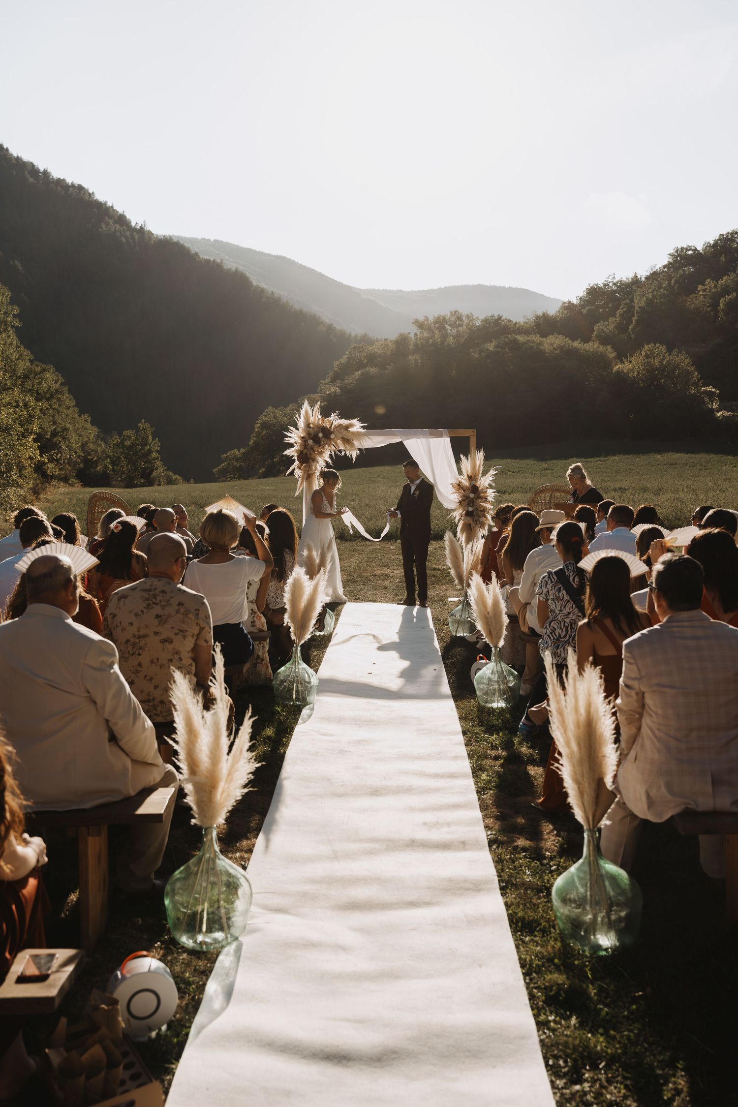Guests seated at outdoor boho wedding ceremony with pampas grass aisle and mountain backdrop.