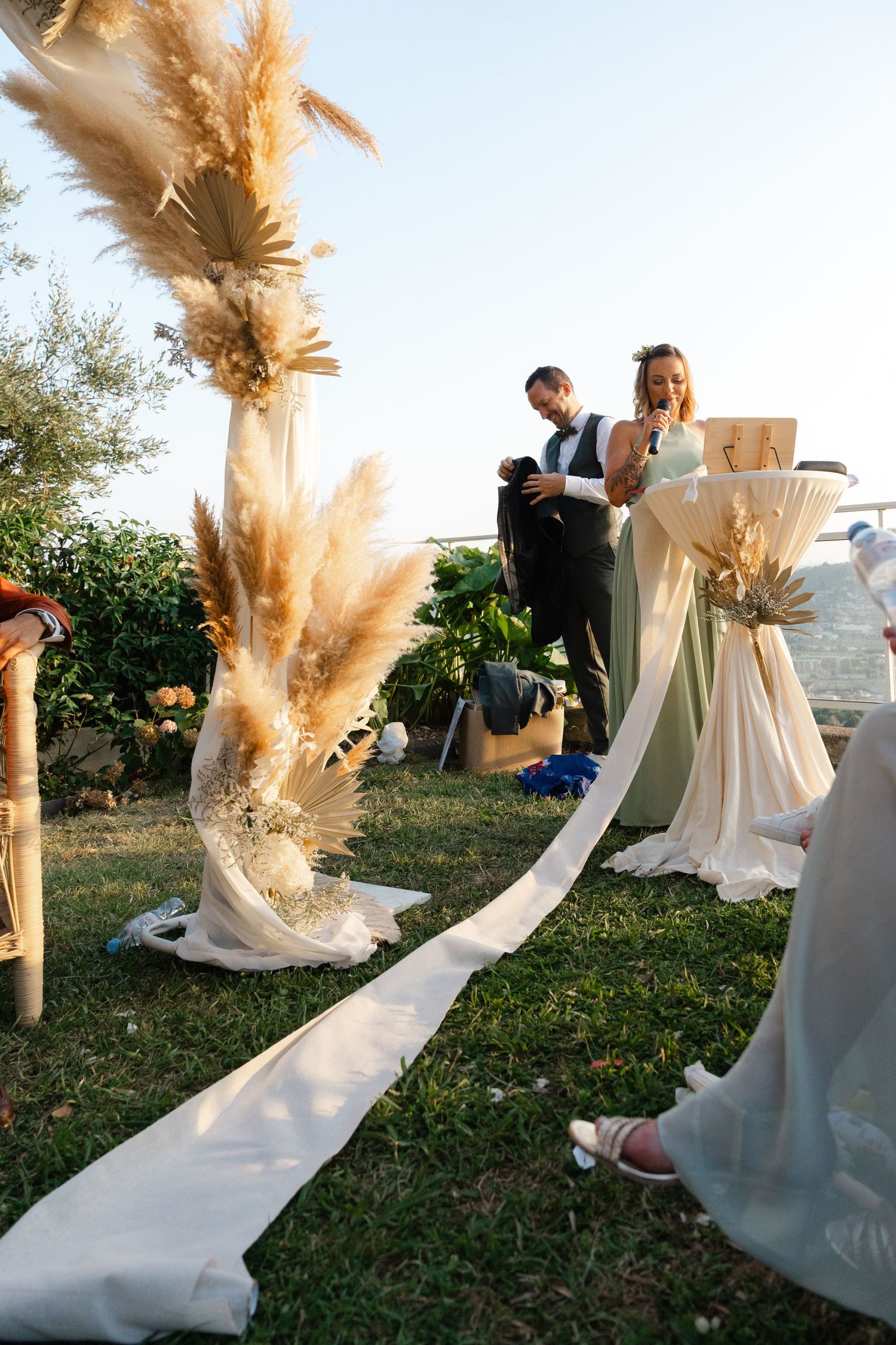 Outdoor wedding ceremony with pampas grass decorations and white fabric runner on green lawn.