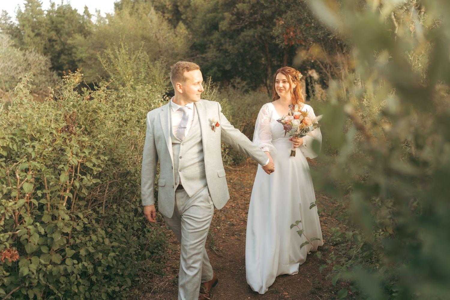 A bride and groom walk hand in hand through a lush garden path on their wedding day.