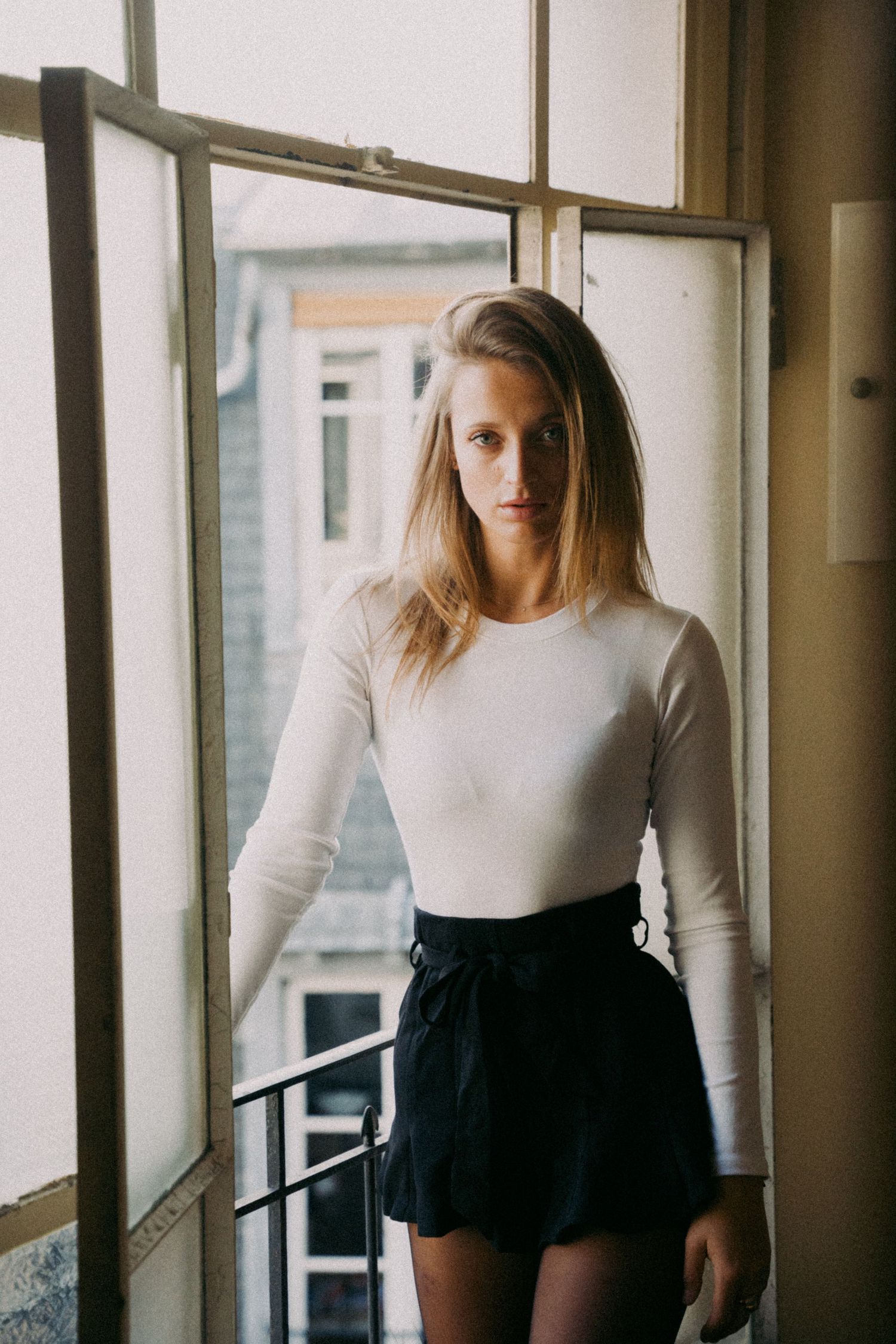 Young blonde woman in a white long-sleeve top and black mini skirt standing by an open window.