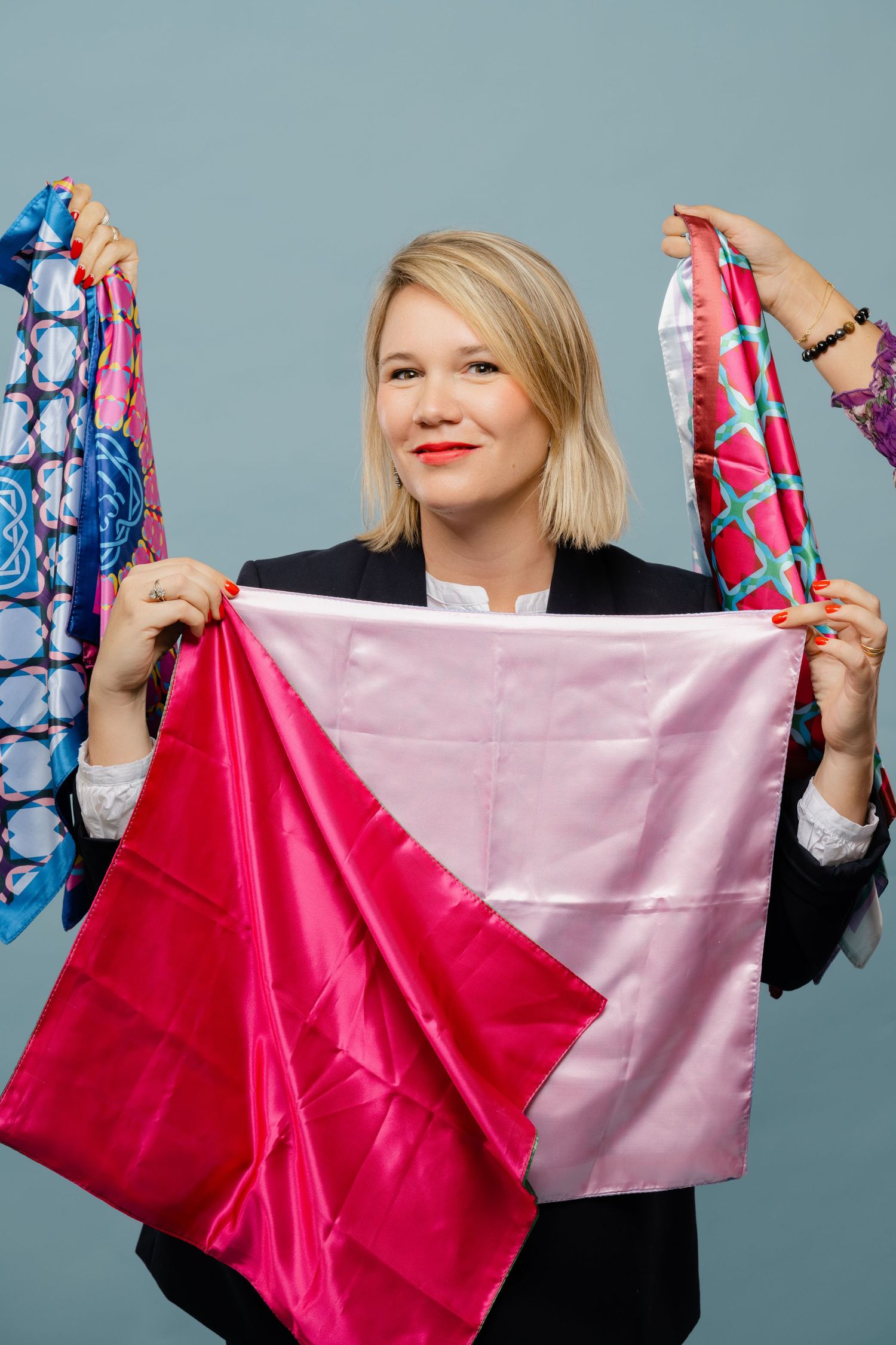 Blonde woman in black blazer holding colorful silk scarves against blue background.