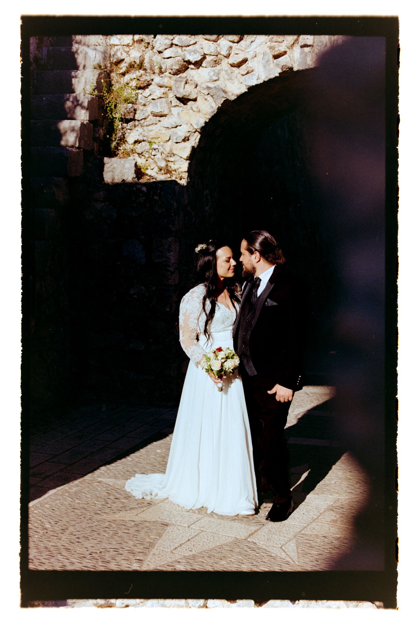 Bride and groom sharing romantic moment under stone arch in vintage film photography style. Shot on film