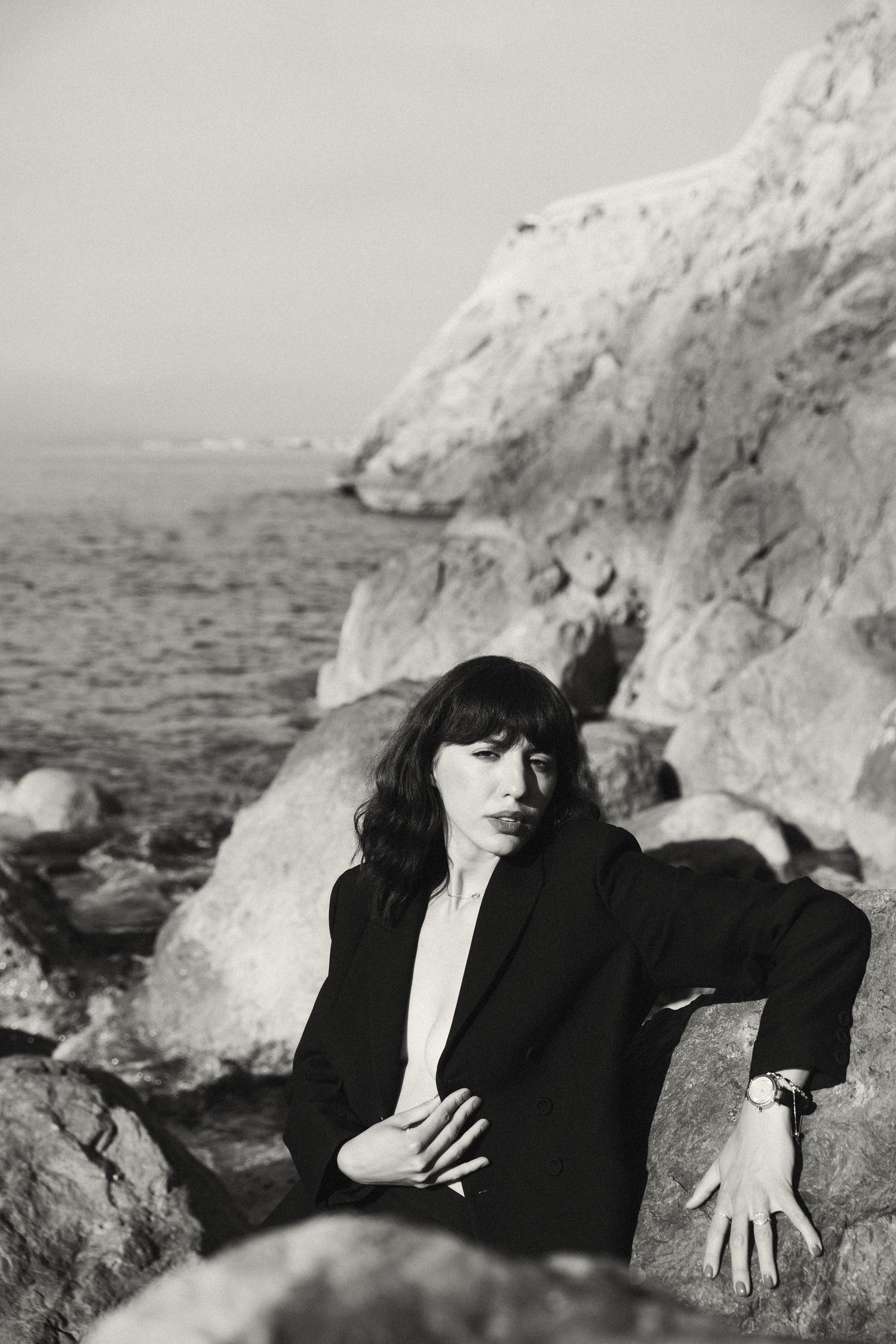 Woman in black blazer poses on coastal rocks in black and white portrait photography.