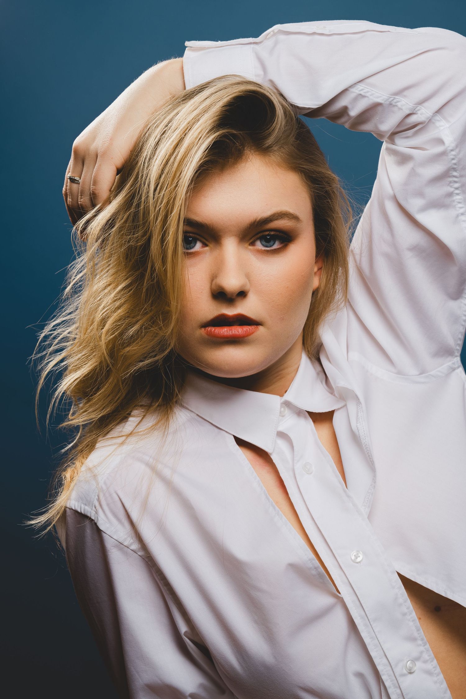 Blonde woman in white button-up shirt posing with hand in hair against dark blue studio background.